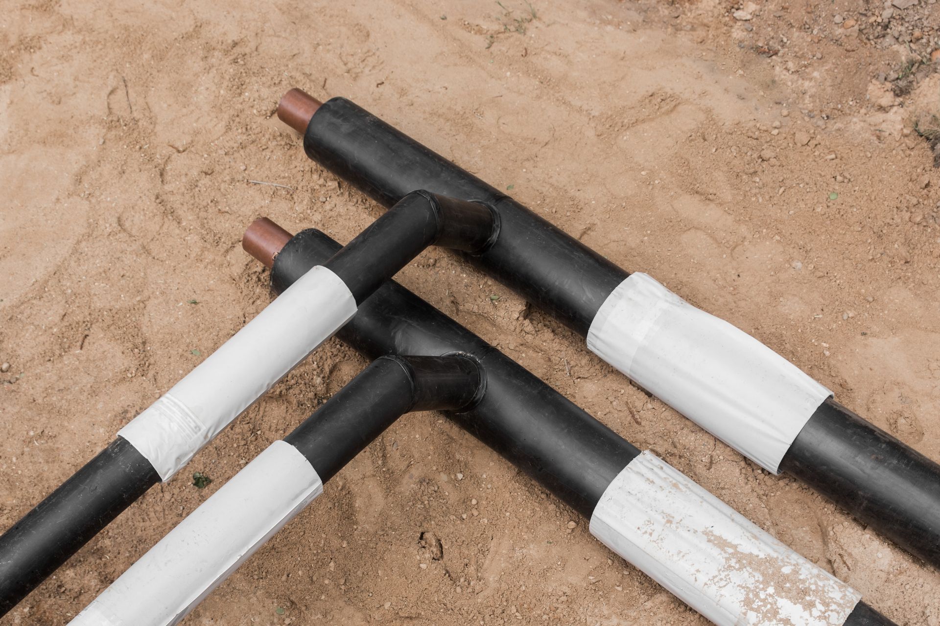 Black and white insulated pipes laid on sand. Two pipes form a T-shape, wrapped in white tape.