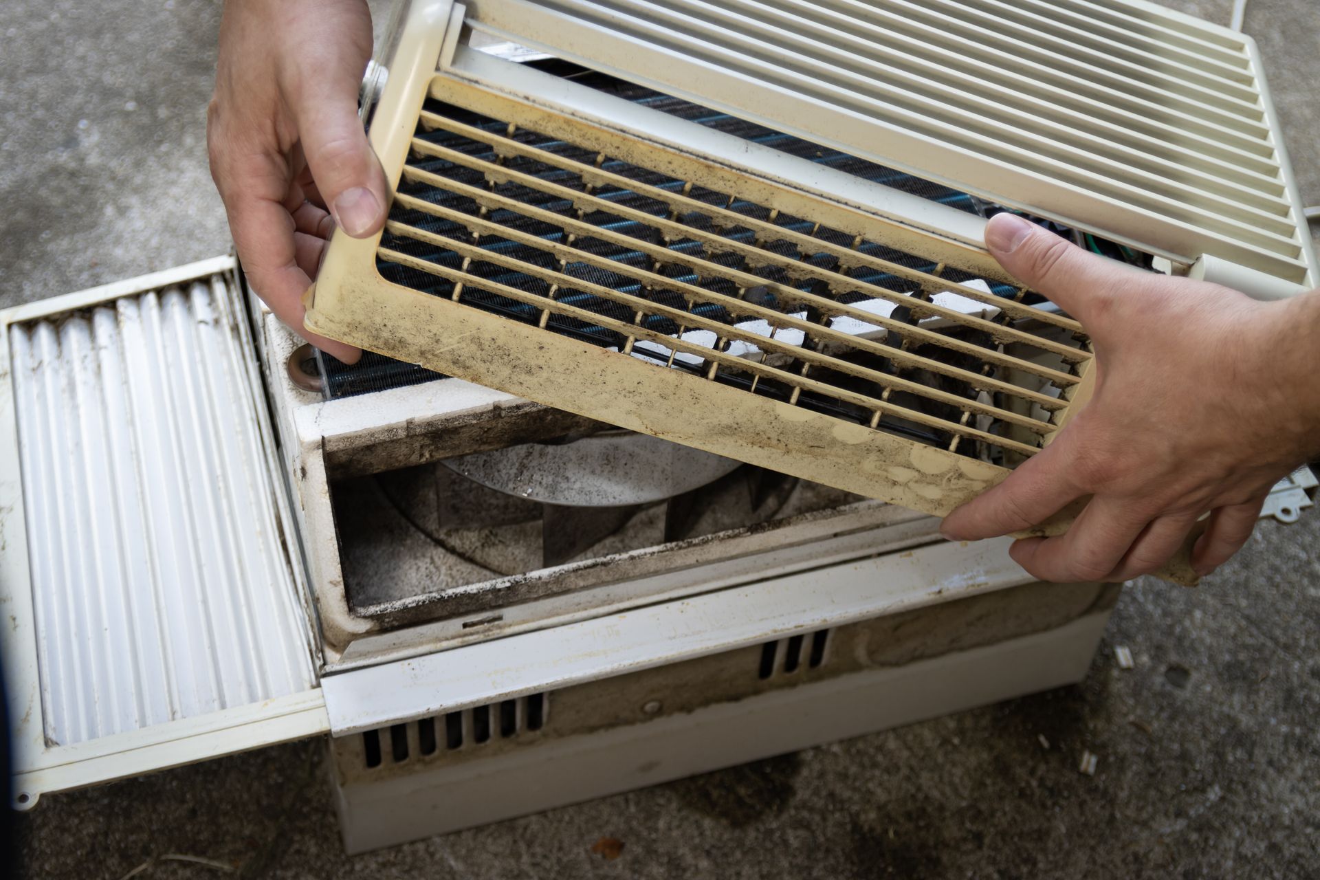 Hands holding a dirty air conditioner vent cover. Unit is open, likely for cleaning.