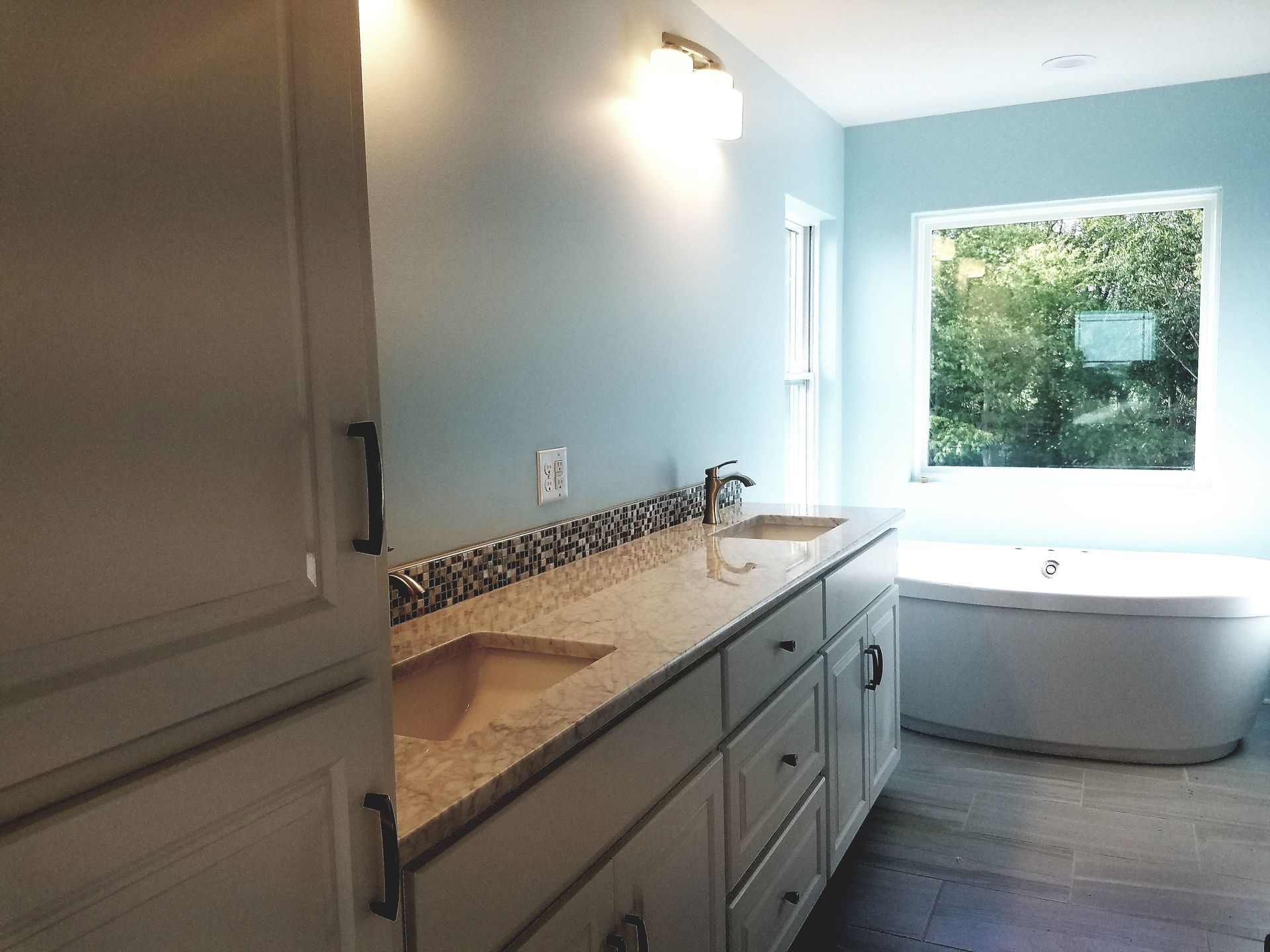Bathroom with light blue walls, a light stone countertop vanity with a sink, and a white soaking tub near a window.