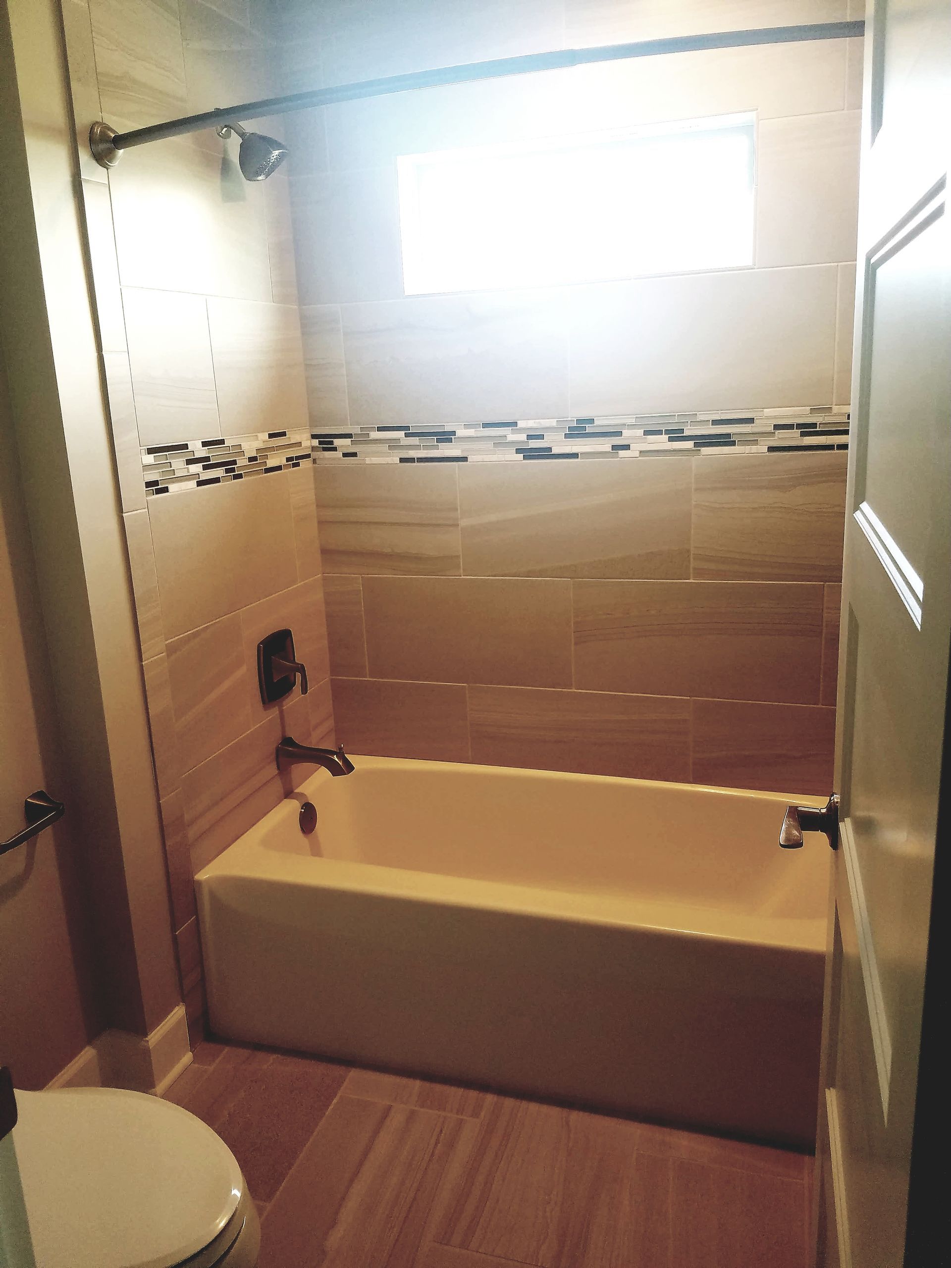 A bathroom featuring a beige bathtub with a shower head, tan tiled walls with a mosaic accent strip, and a white toilet.