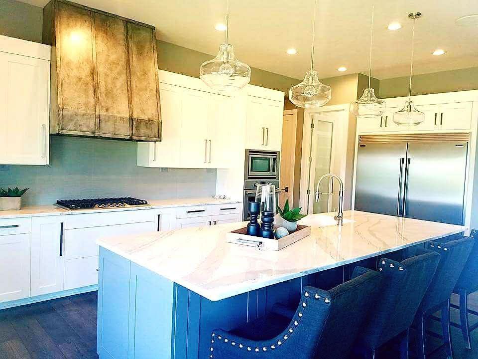 A modern kitchen with a large blue island, white cabinets, stainless steel appliances, and a rustic metal range hood.