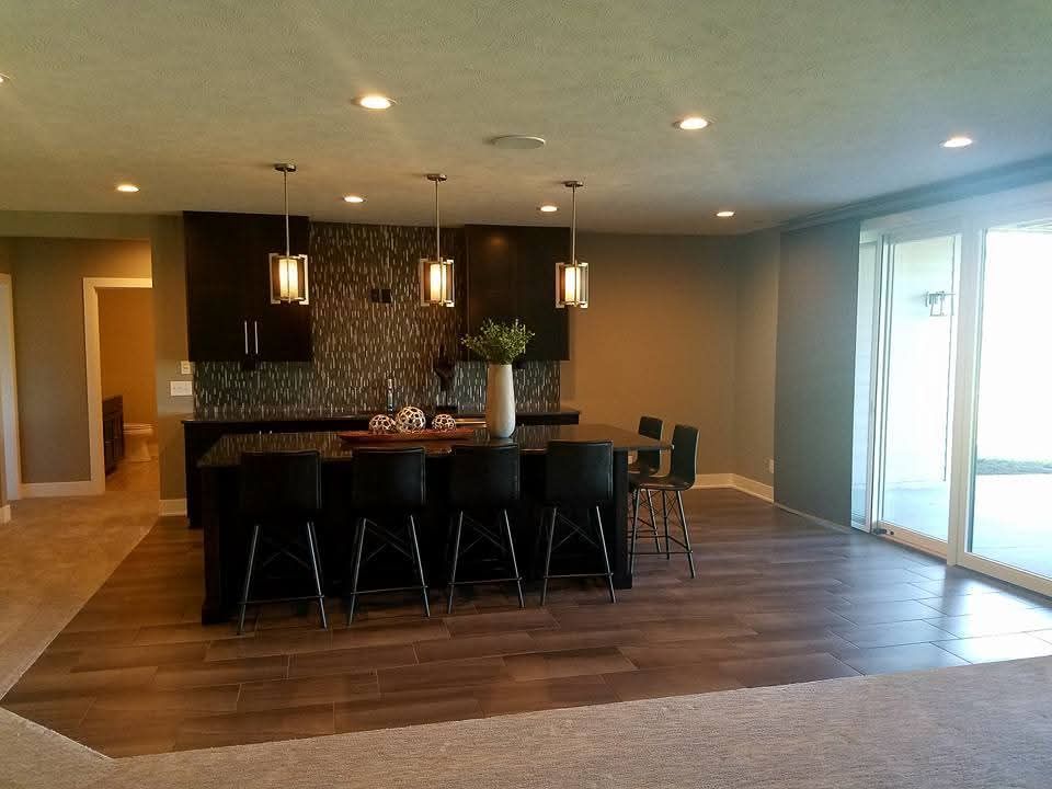 Modern kitchen with a dark wood island, bar stools, pendant lighting, and dark cabinets against a textured backsplash.
