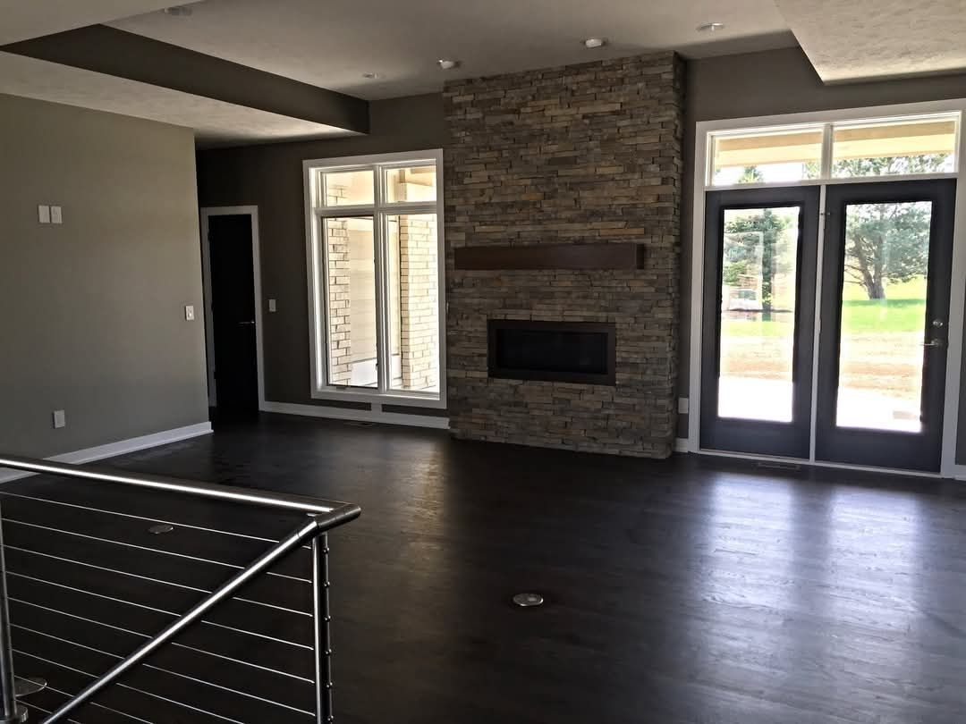 A modern living room with dark wood floors, a stone fireplace, large windows, and a glass-railed staircase.