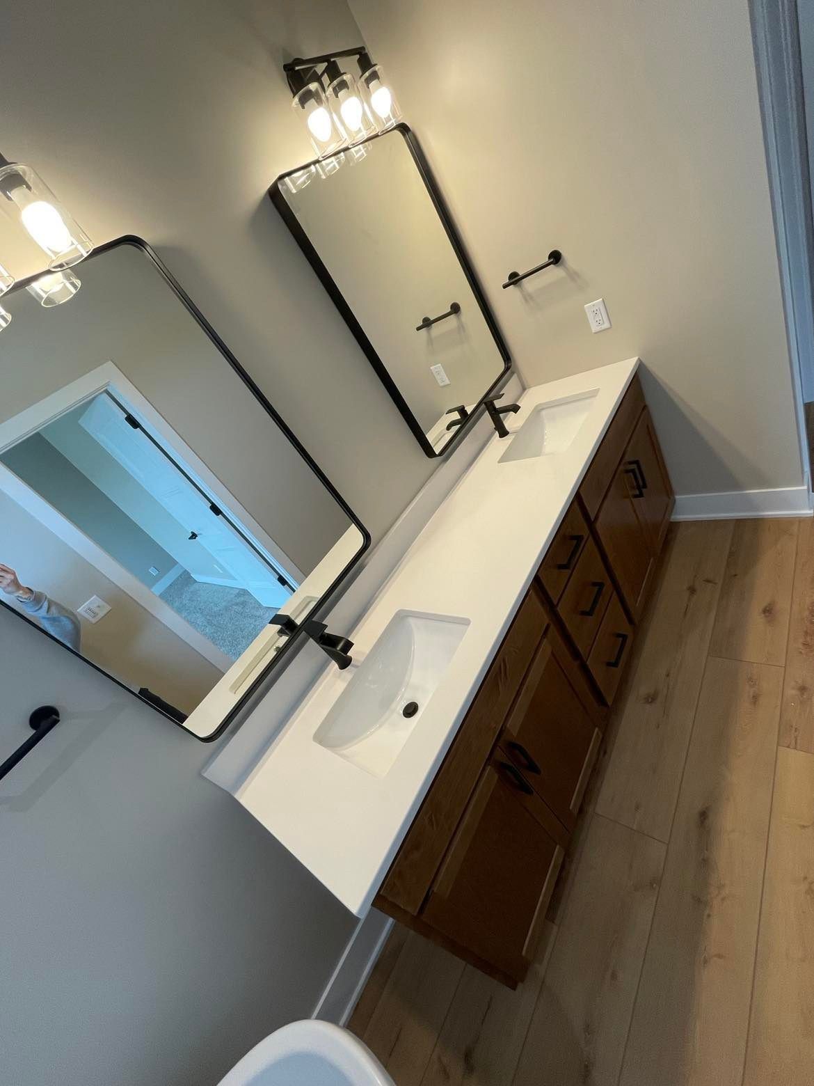 A modern bathroom featuring a double vanity with white countertops, wooden cabinets, two framed mirrors, and light fixtures.
