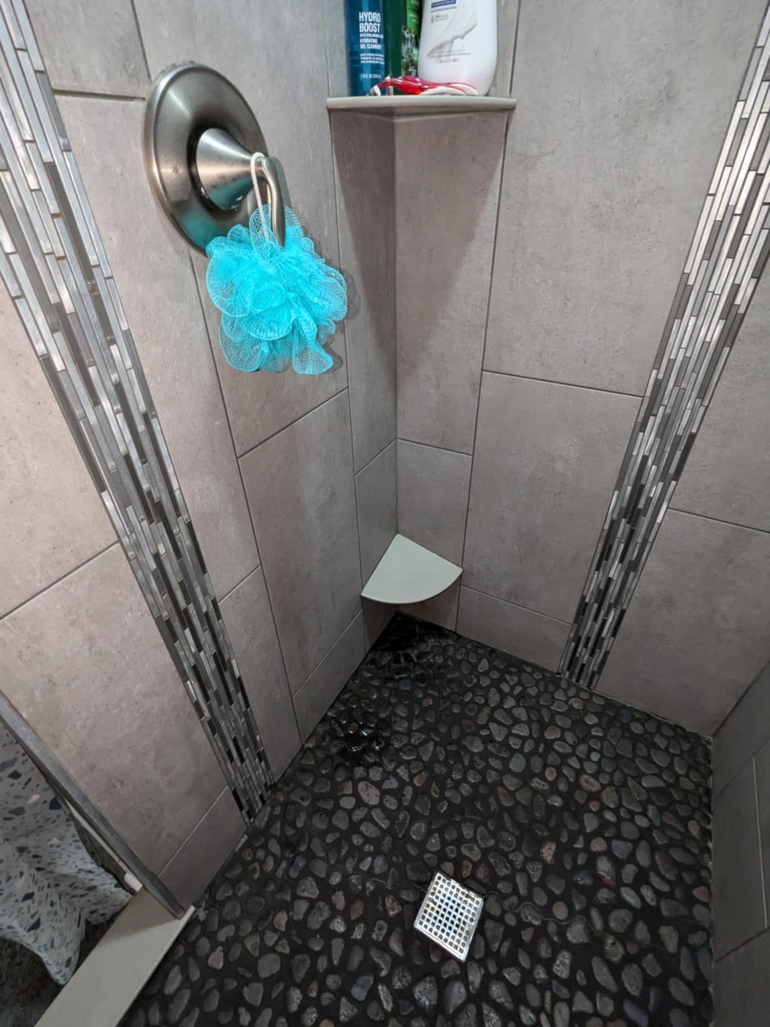A shower corner with gray tiled walls, decorative vertical stone accents, a corner shelf, and a blue mesh sponge on faucet.