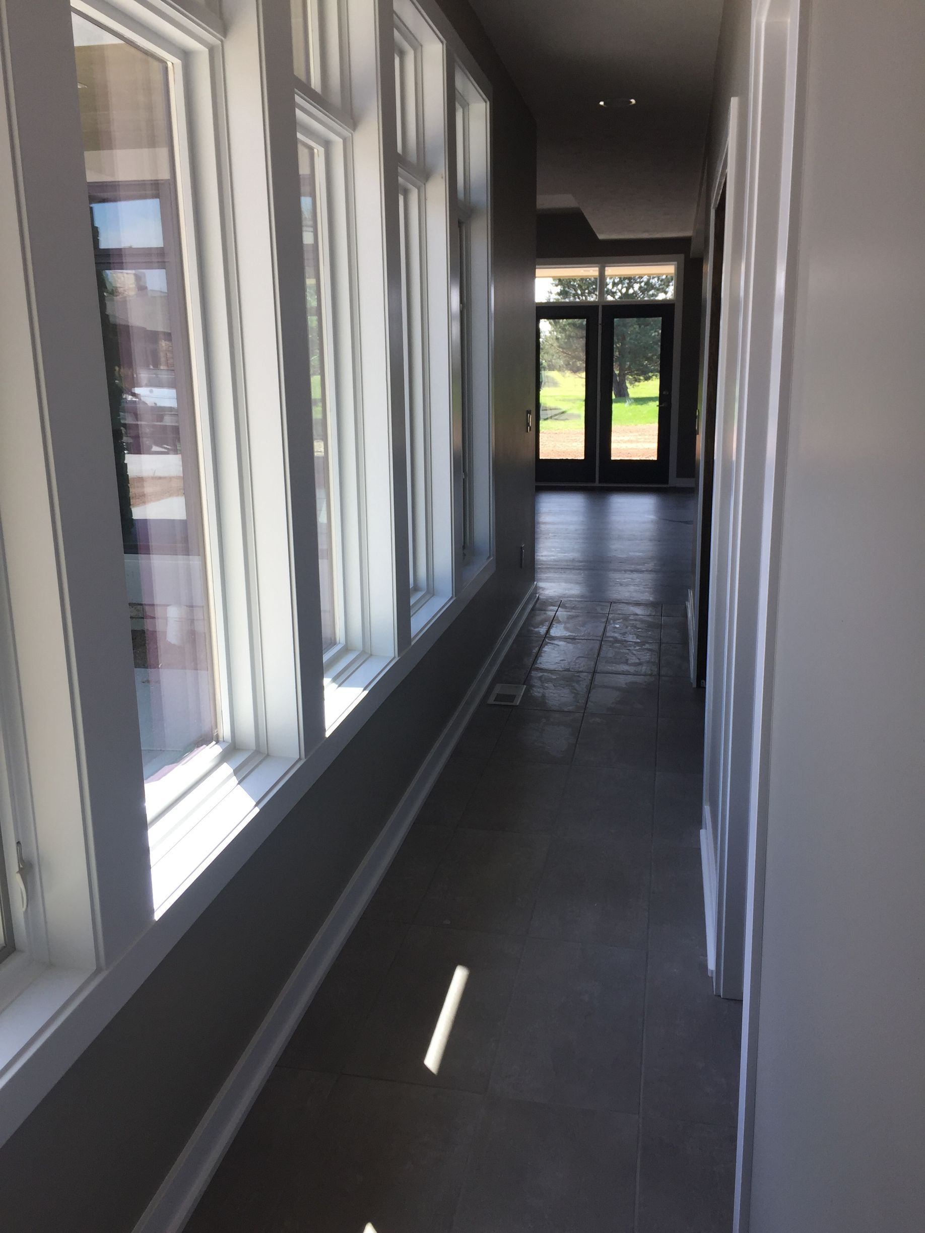 A long indoor hallway with grey tile flooring, lined with a series of tall windows, leading toward glass double doors.