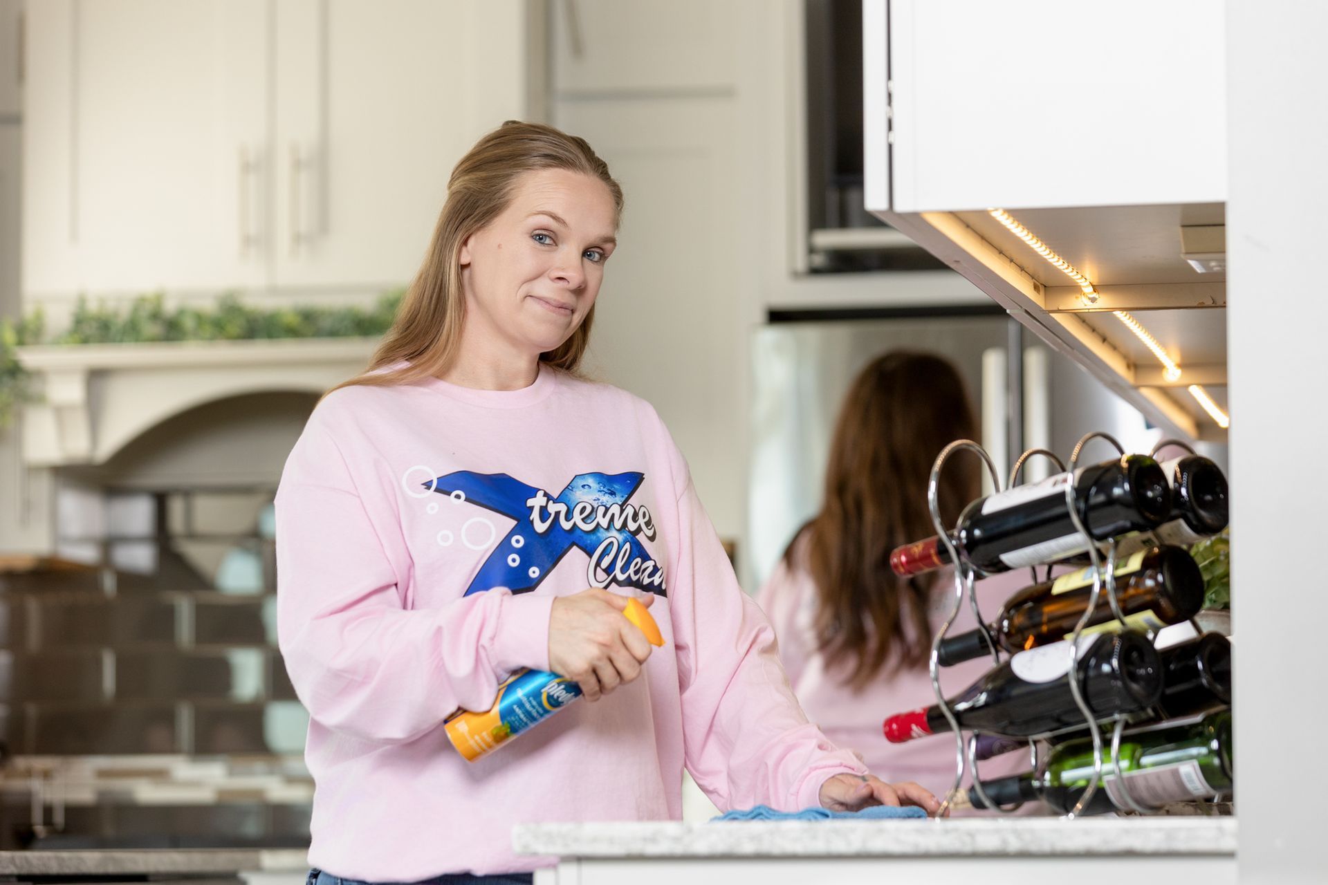 A person in a pink sweater holds a spray bottle while cleaning a kitchen counter near a wine rack.