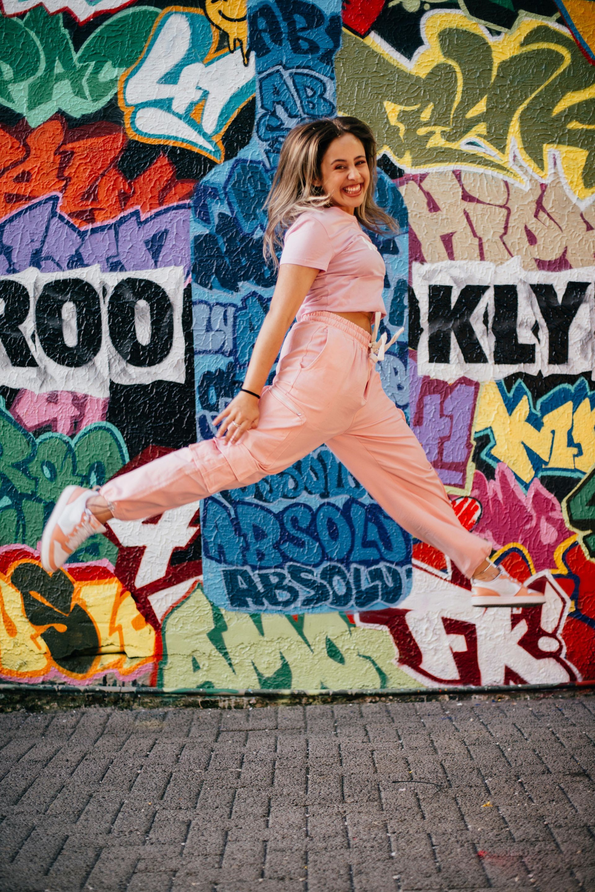 Woman in pink jumpsuit jumps mid-air in front of graffiti-covered wall, smiling.