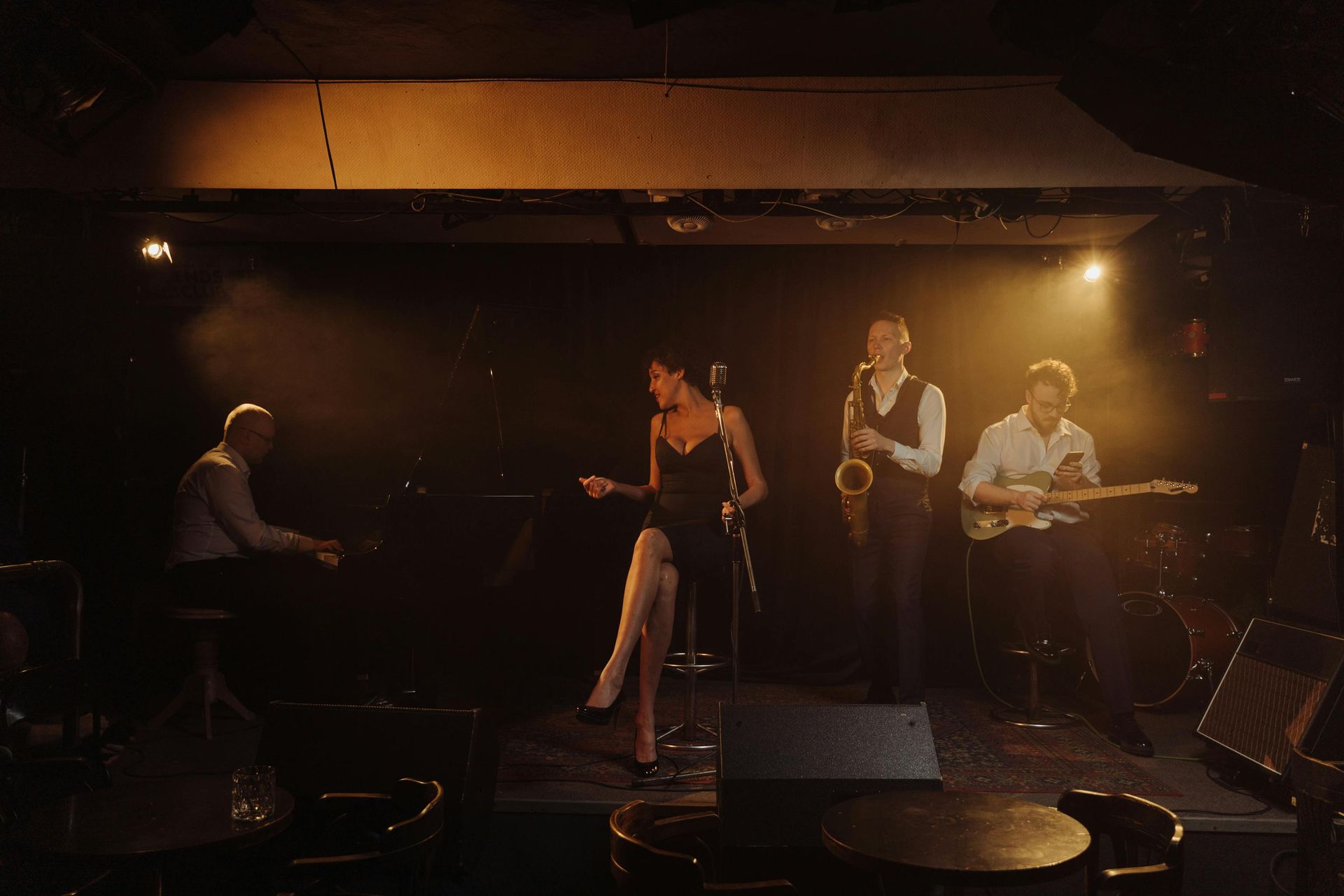 Jazz band performing on a dimly lit stage: singer, piano, sax, and guitar, bathed in warm spotlights.