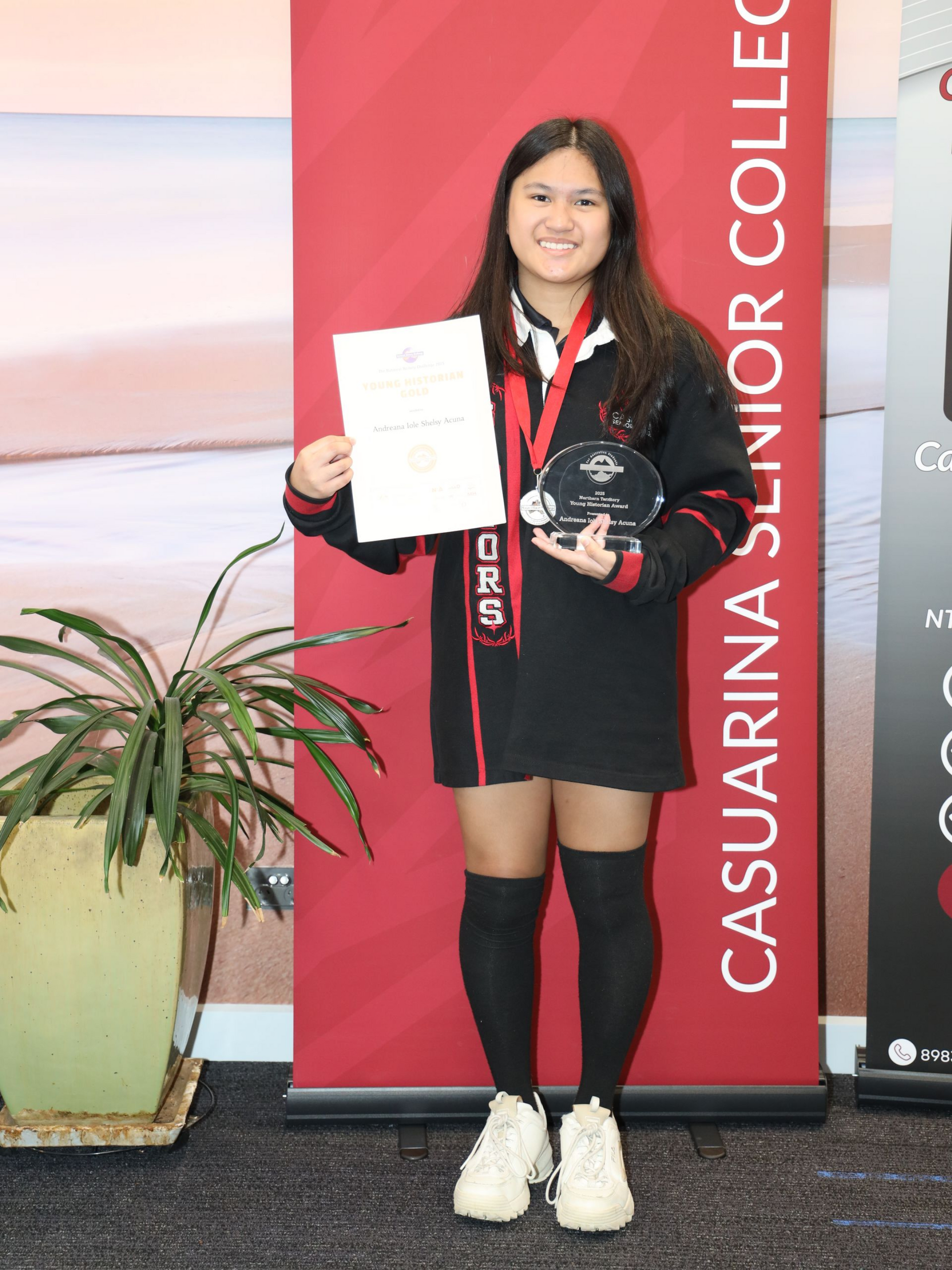 Teenage girl holding award and certificate, standing in front of school banner, smiling.