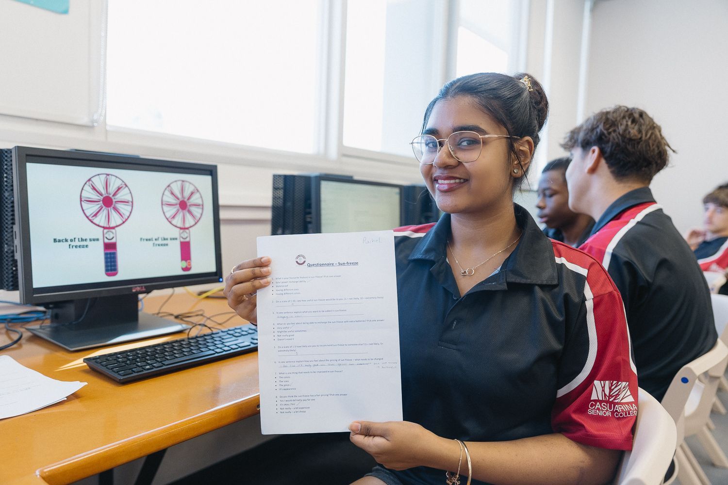 Woman in a school uniform holds a paper in a computer lab, smiling. A diagram is on the computer screen.
