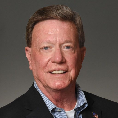Head-and-shoulders portrait of a man in a dark blazer and checkered shirt, smiling against a gray background.