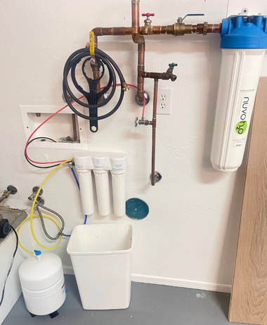 Plumbing wall setup with pipes, valves, water filter, hoses, and utility buckets in a utility room.