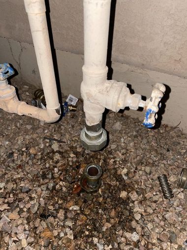 Plumbing pipes and shutoff valves under a sink, with a drain opening in a gravel floor.