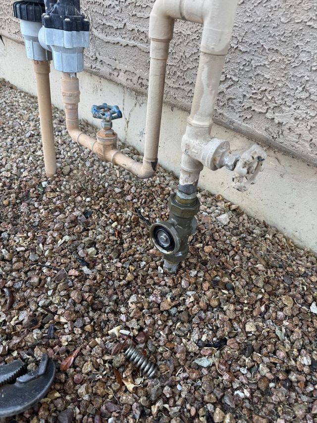 Outdoor water meter and pipes beside a stucco wall over gravel.