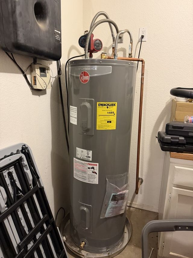 Gray water heater with pipes and labels in a utility closet, beside a black wall unit and stacked items.