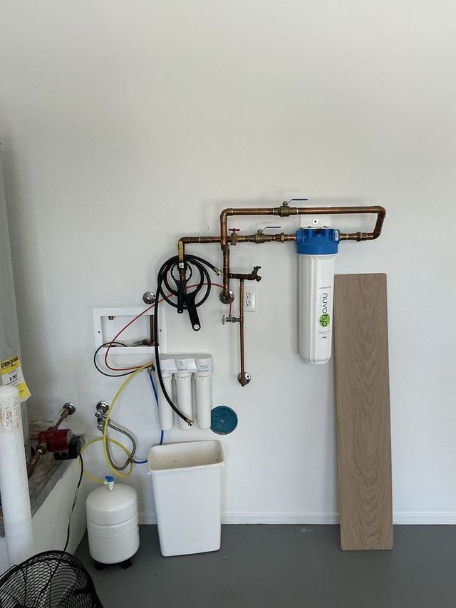 Wall-mounted water filtration system with white tank, blue filter, and pipes against a white wall