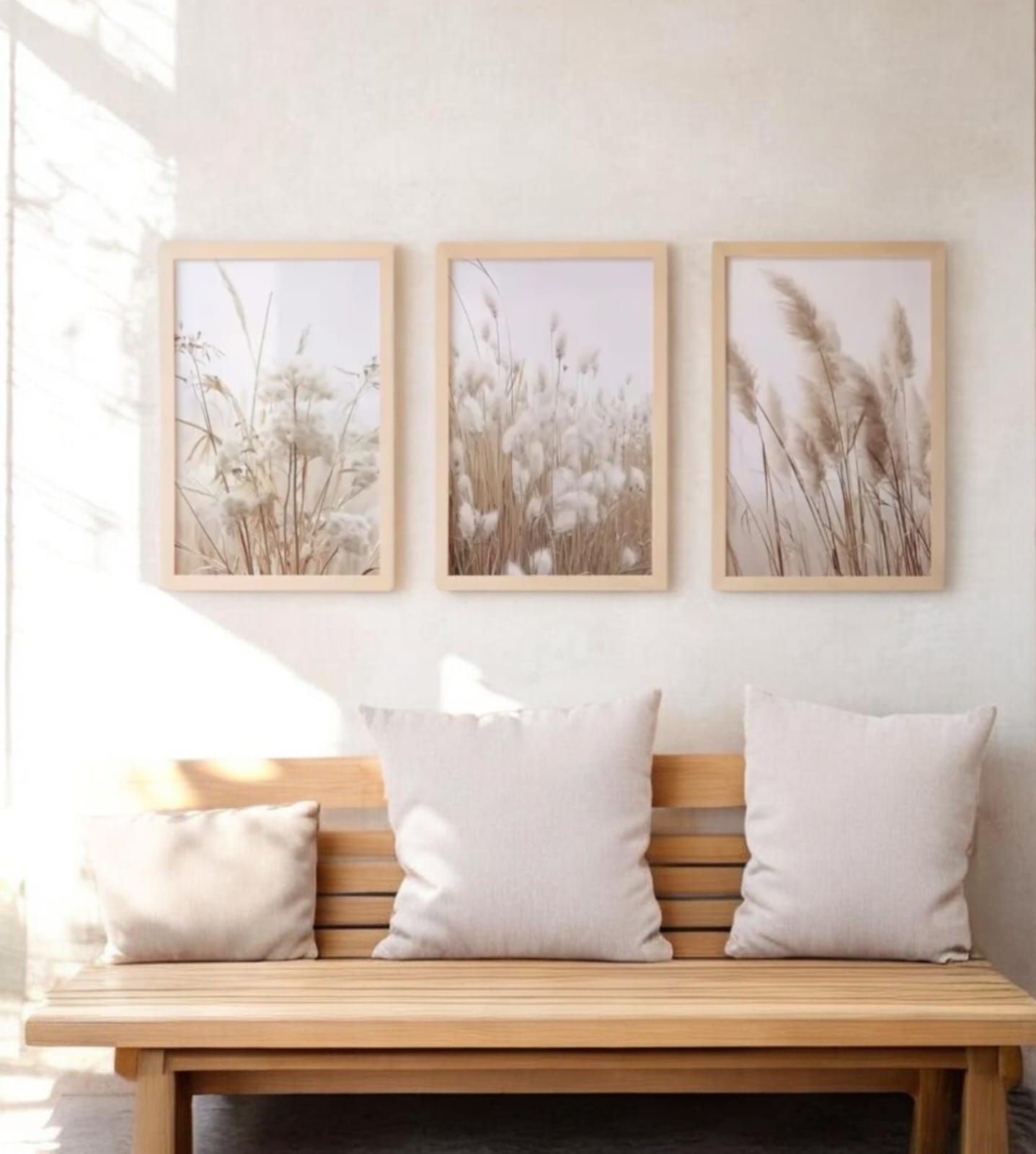 Banco minimalista de madera con cojines blancos y tres láminas botánicas enmarcadas de color beige sobre una pared clara.