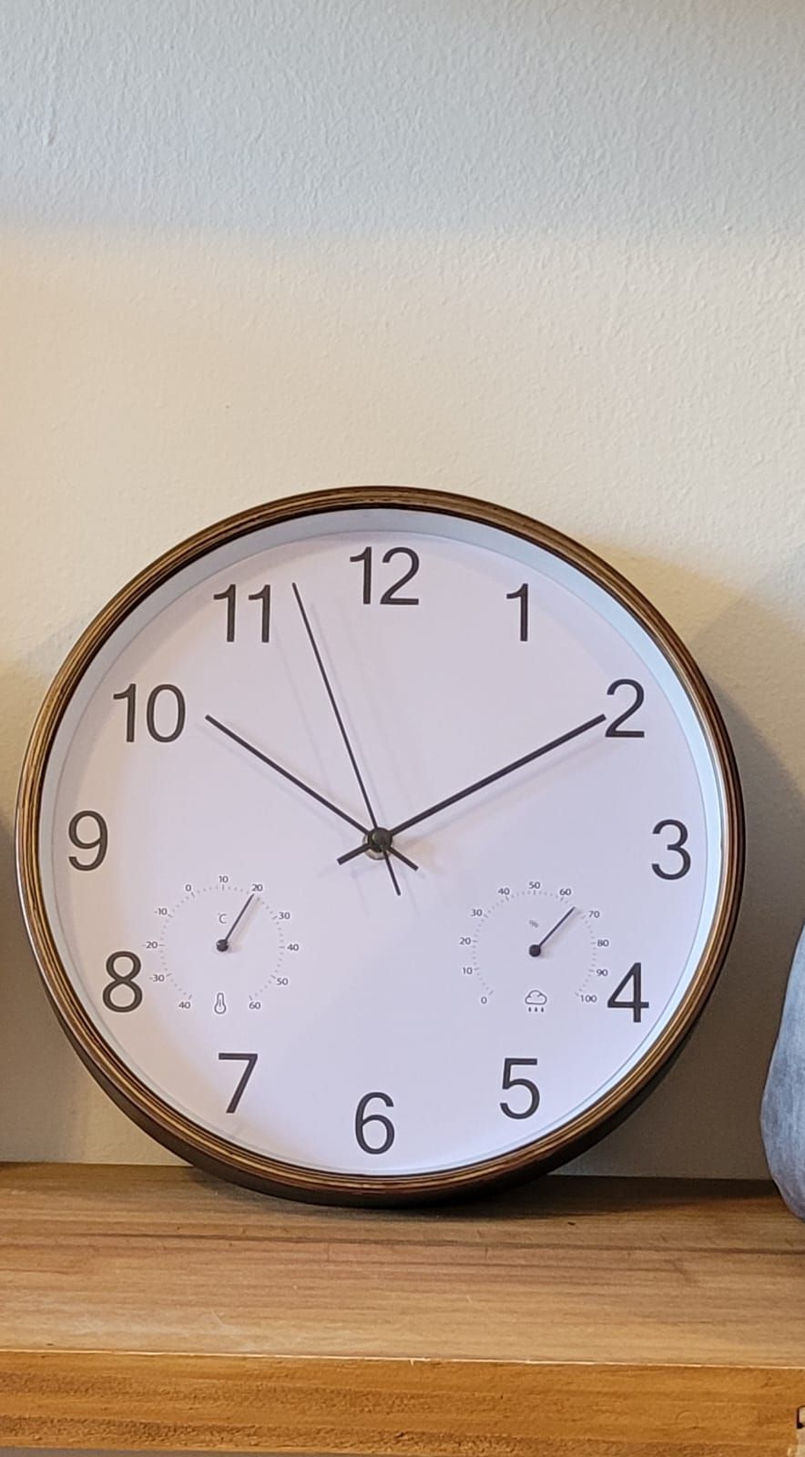Reloj de pared redondo de madera sobre una repisa, que marca aproximadamente las 10:10 sobre una pared blanca.
