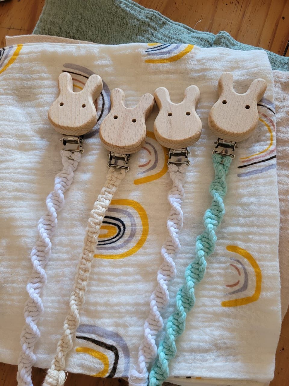 Cuatro clips para chupetes con forma de conejito en color blanco, crema y turquesa sobre una tela estampada.