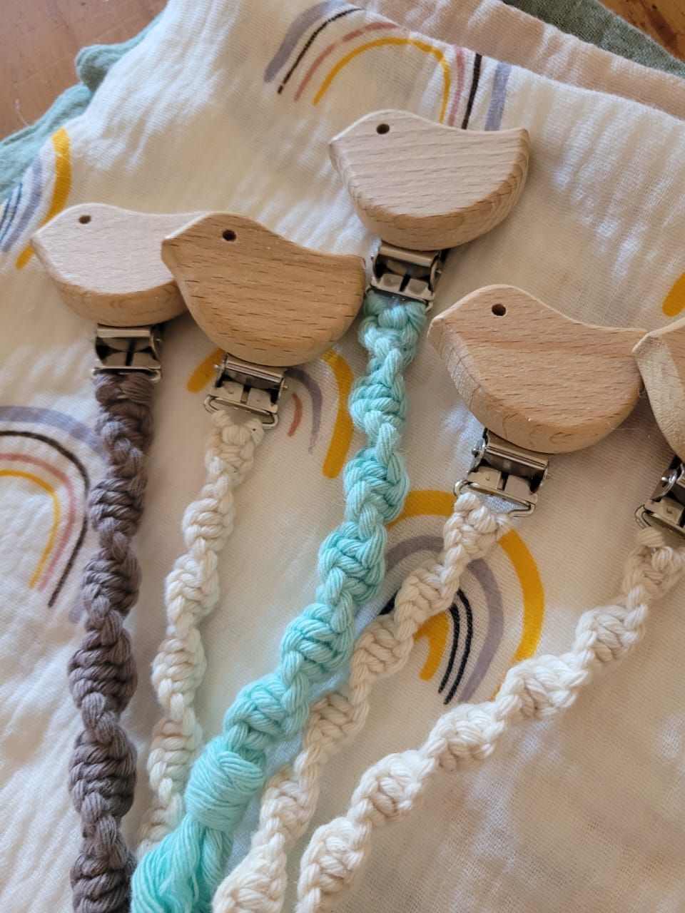 Cuatro pinzas de madera con forma de pájaro en una tela a rayas color crema, una de ellas con un cordón trenzado turquesa.