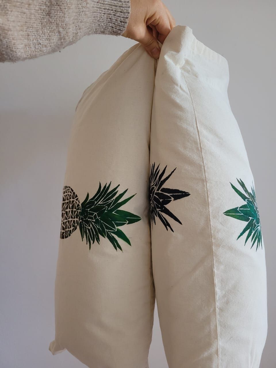Una mano sostiene dos almohadas color crema con estampados de hojas verdes sobre un fondo claro.