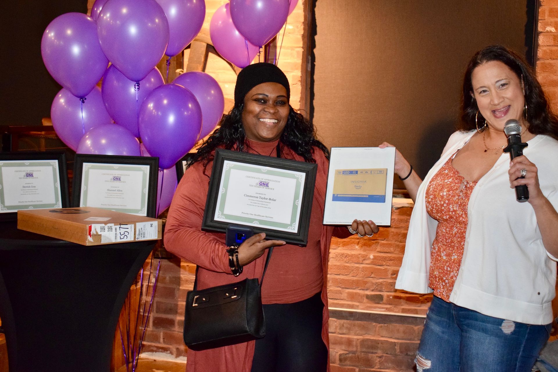 Two people holding framed awards beside purple balloons at a celebration event