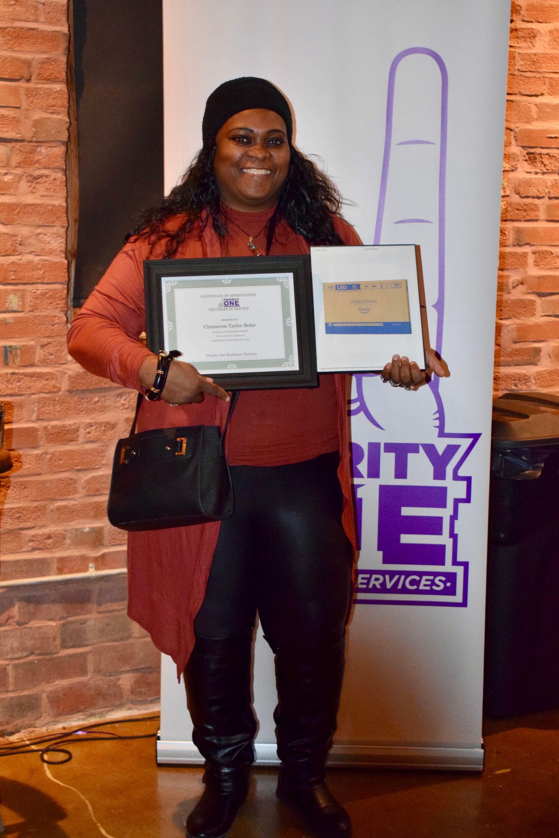 Woman holding two framed certificates in front of a branded banner, smiling indoors.
