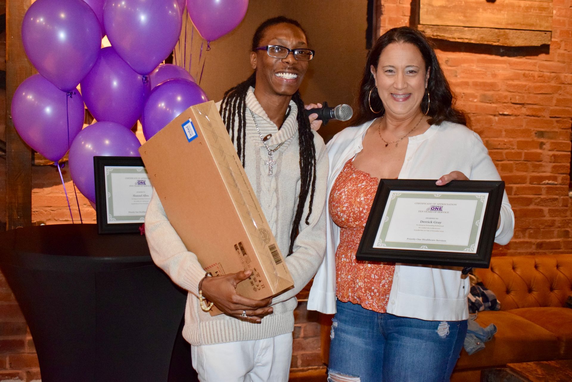 Two people posing with framed certificates and a large box beside purple balloons at an event.