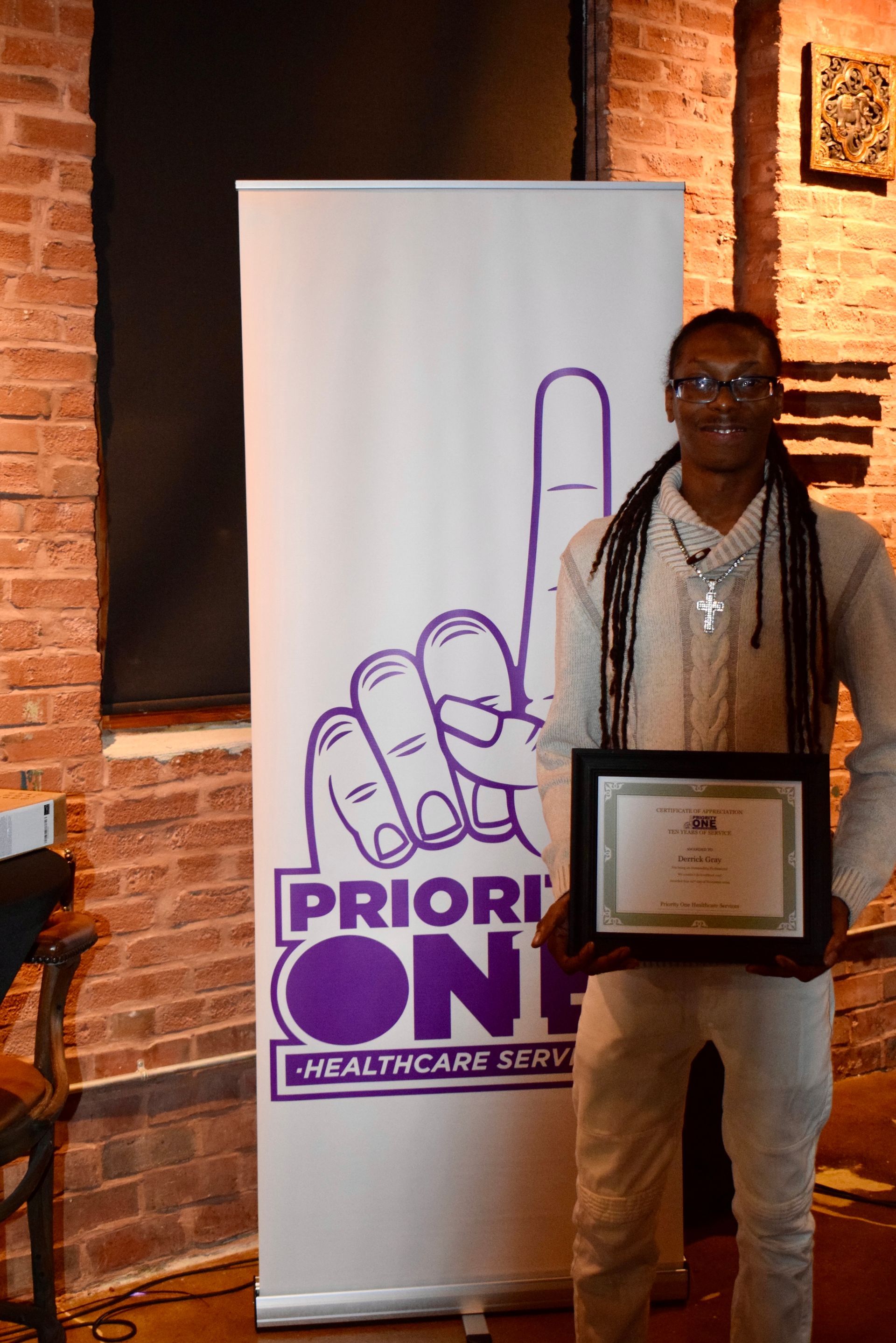 Person holding a framed certificate beside a PriorityON banner in a brick-walled room