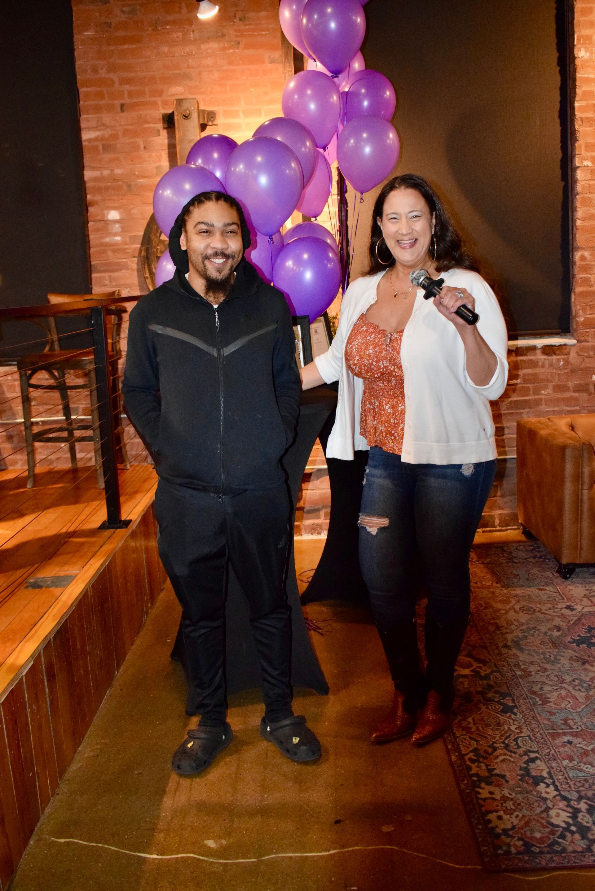 Two people stand indoors by purple balloons, smiling and holding a microphone.