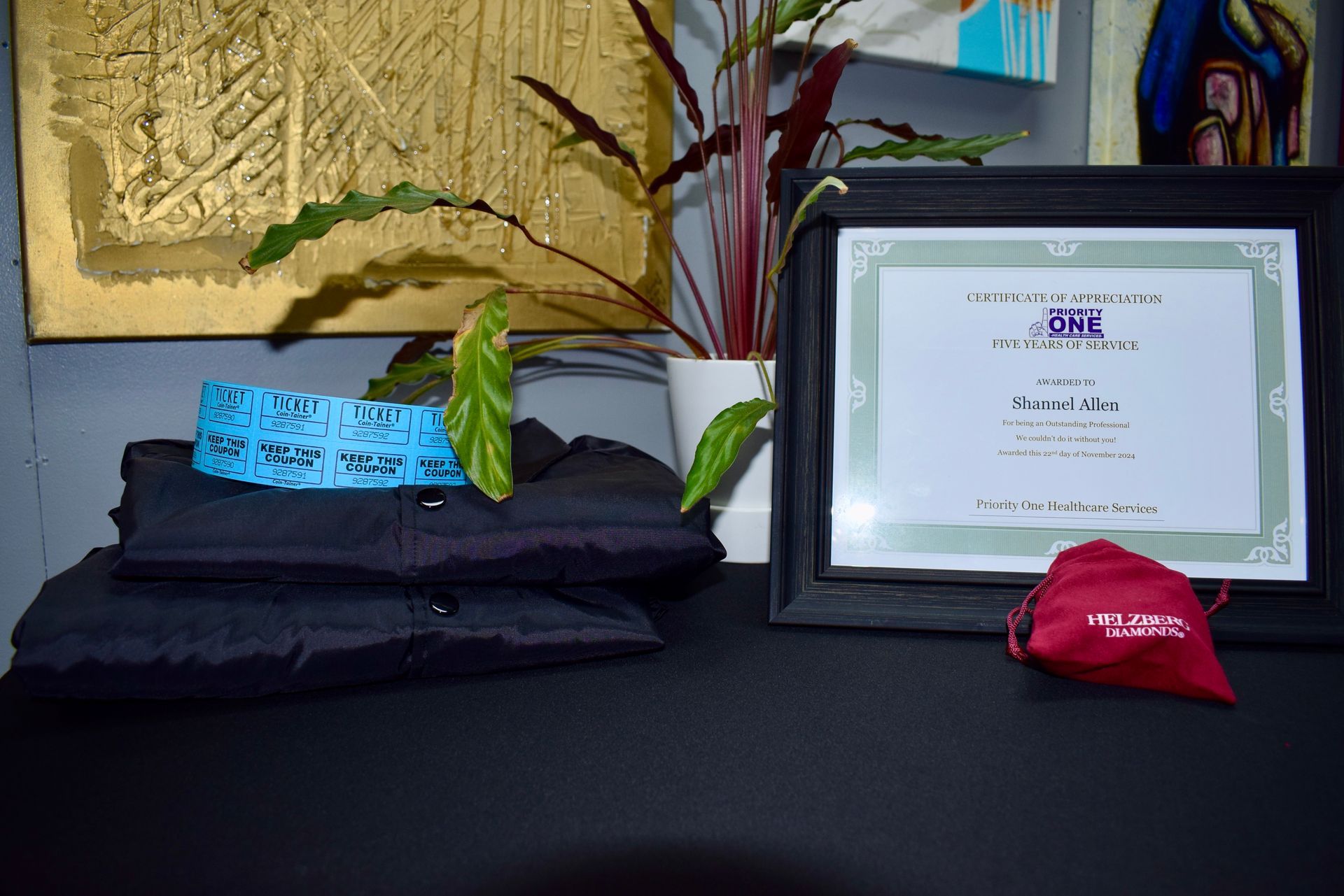 Framed certificate beside folded black shirts, blue and red badges, and a potted plant on a table