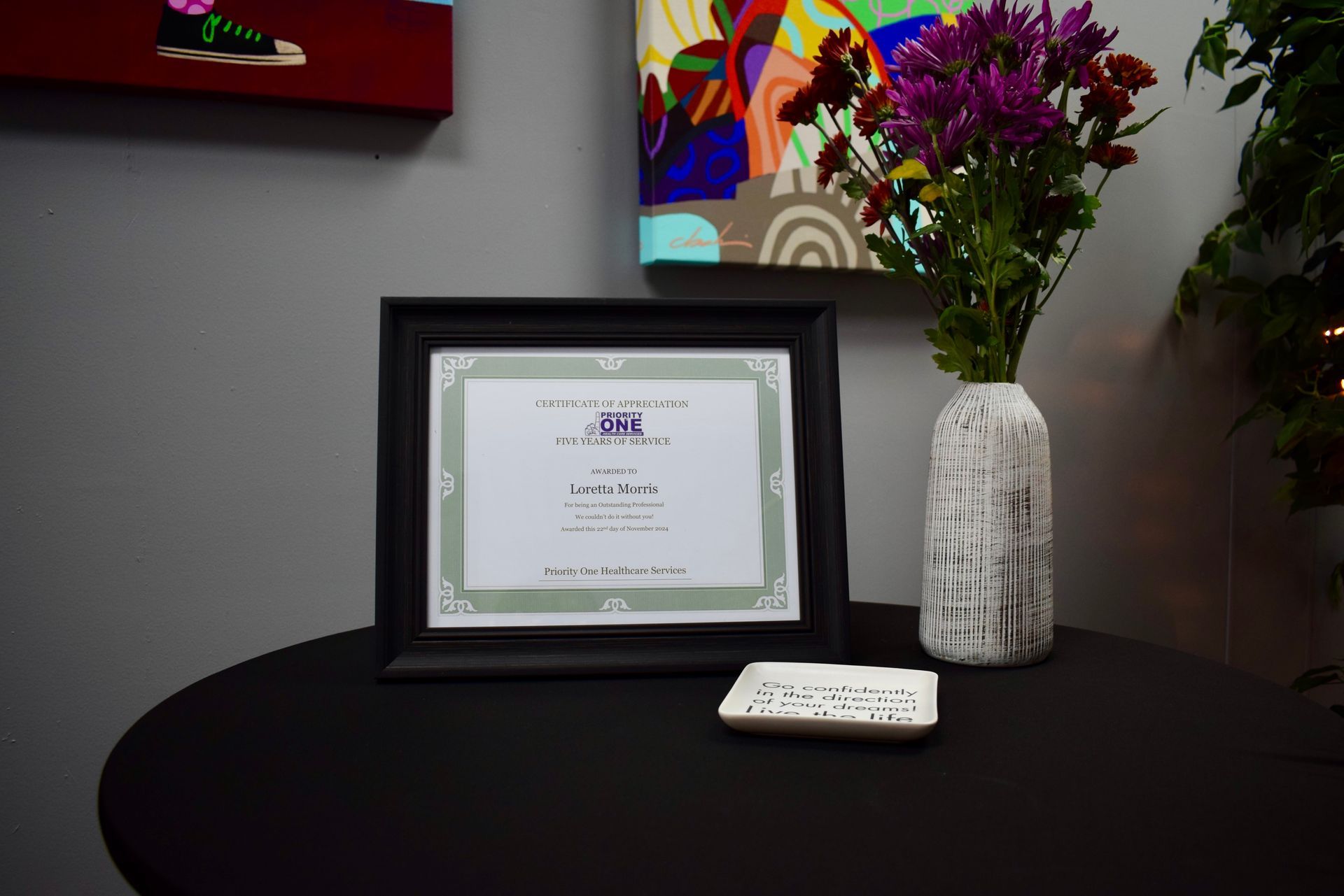 Framed certificate on a black table beside a white vase of purple flowers and a remote control.
