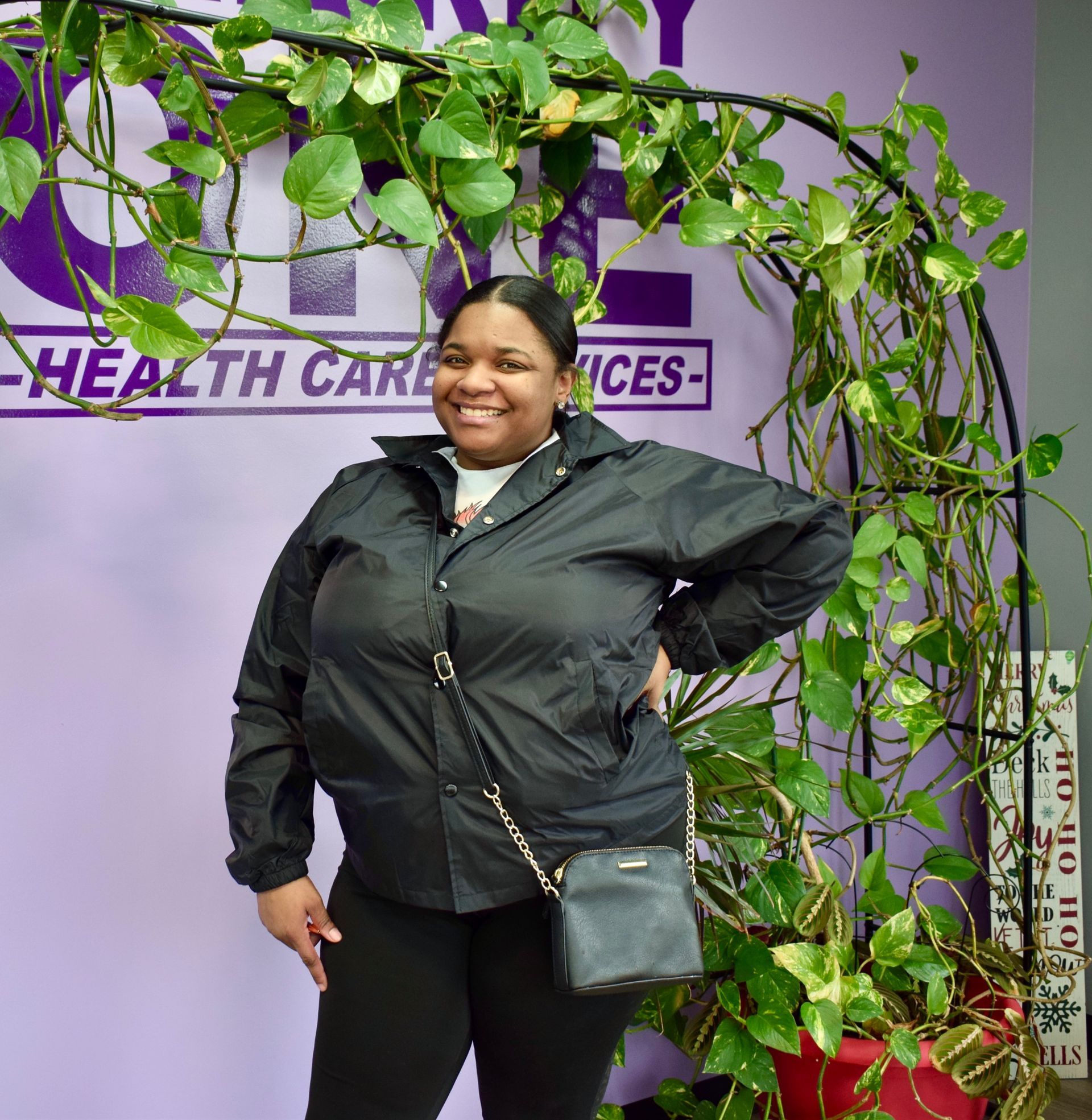 Woman smiling in a black jacket, standing by a leafy plant wall and health services sign.