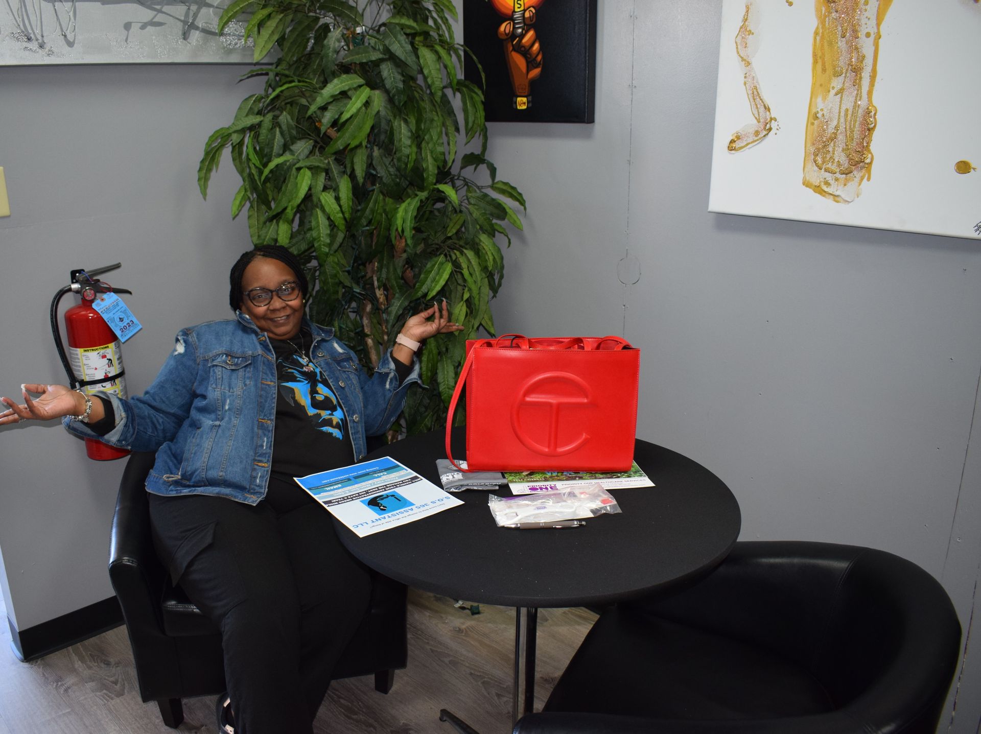 Person seated at a round table, smiling and gesturing in an office with a red bag, papers, plant, and artwork.