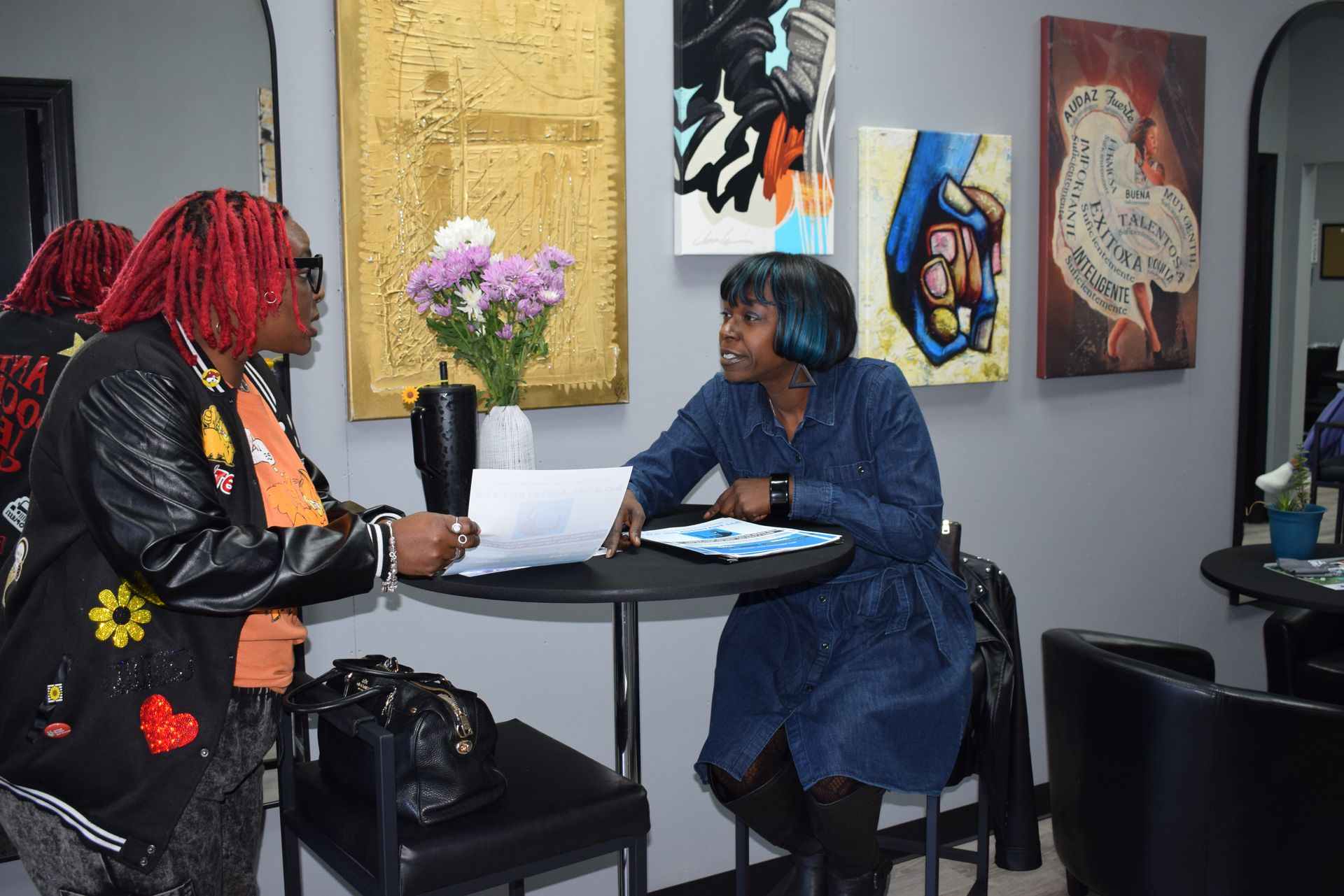 Two people sit at a table in an art gallery, talking over papers with colorful paintings on the wall.
