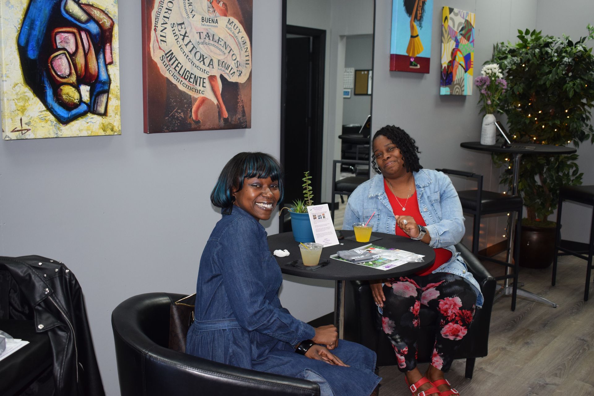 Two people seated at a small round table in a cozy lounge with colorful wall art and plants