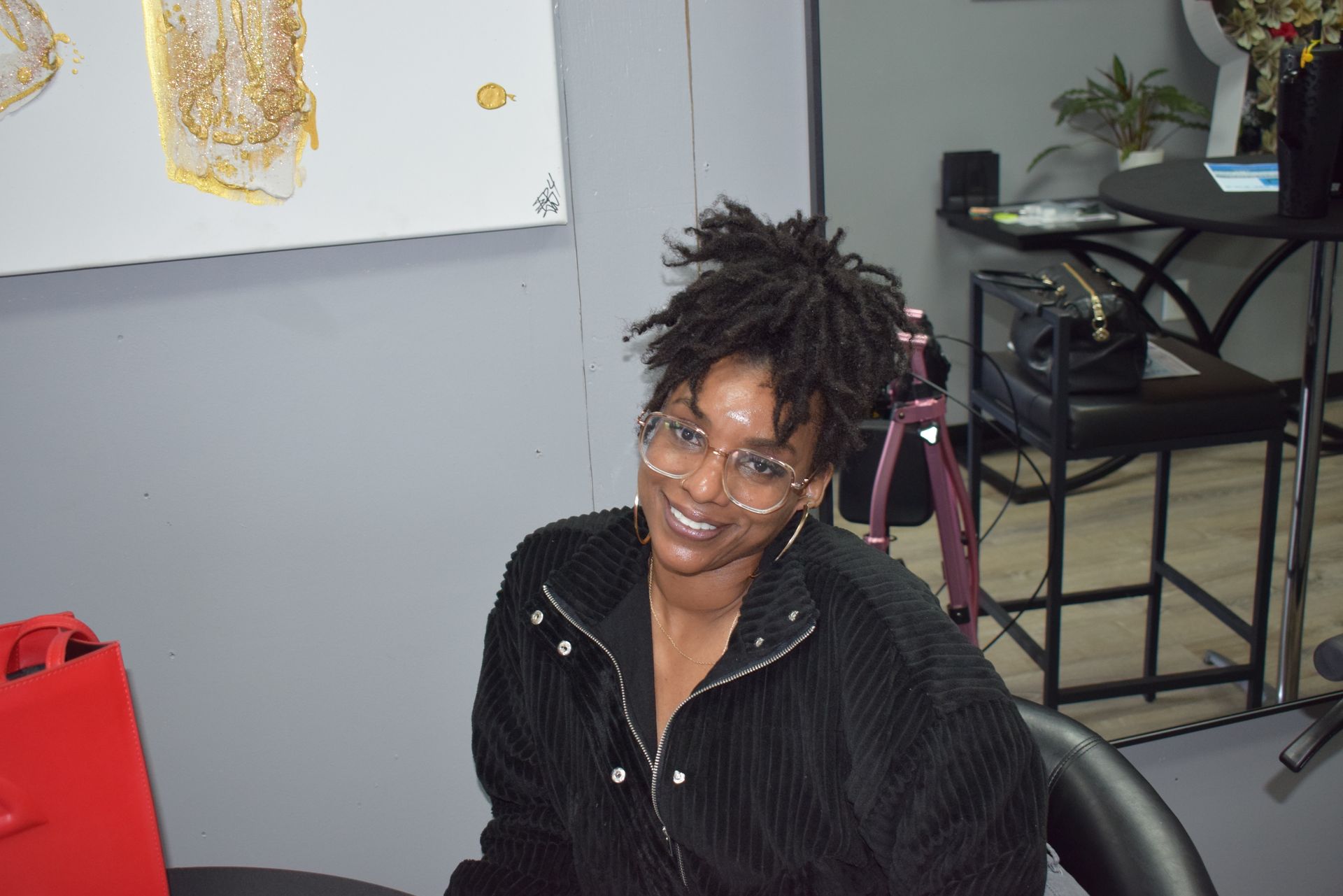 Person smiling in a black jacket, seated in an office near a red bag and desk chairs.