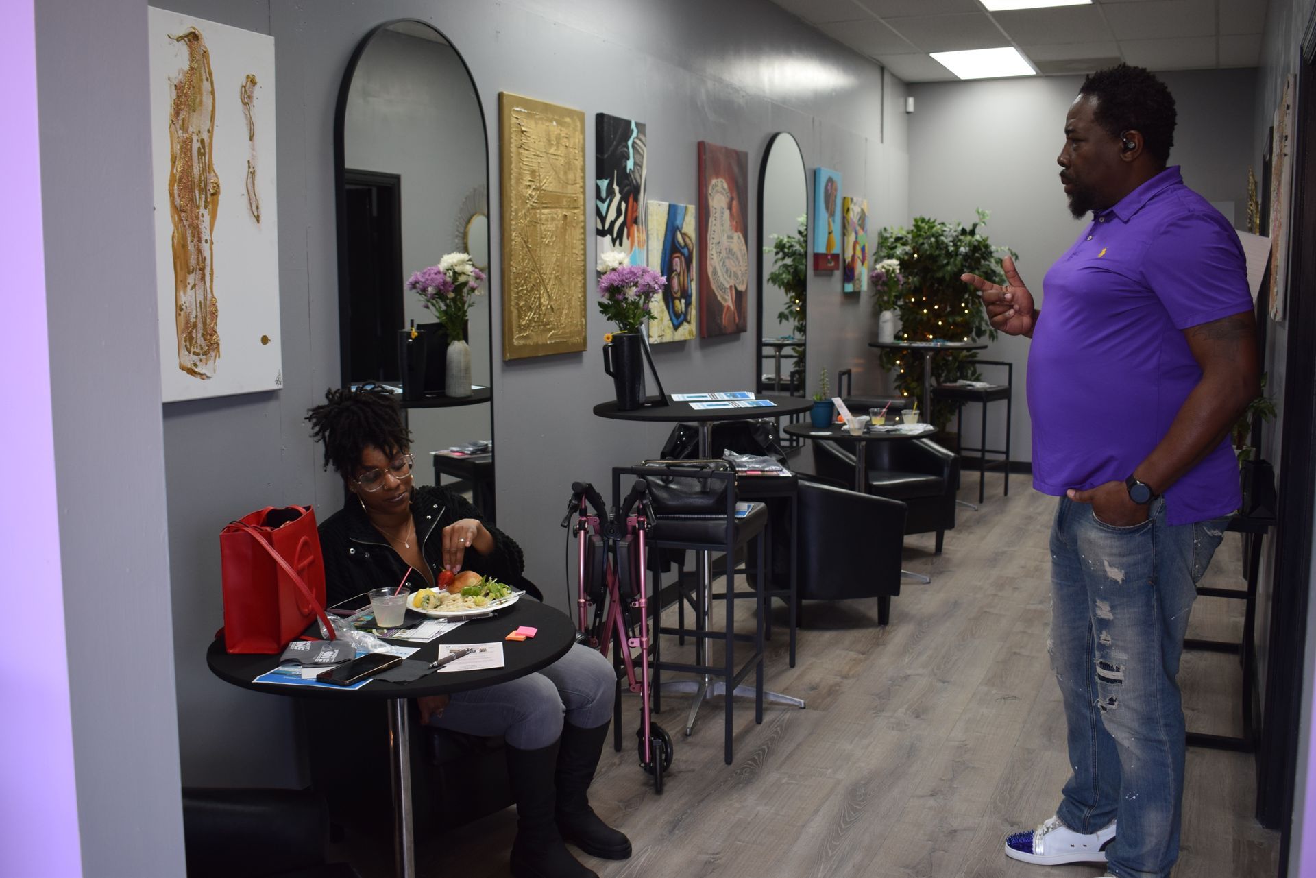 Hallway salon with framed art, a seated person at a small table, and a man standing nearby.