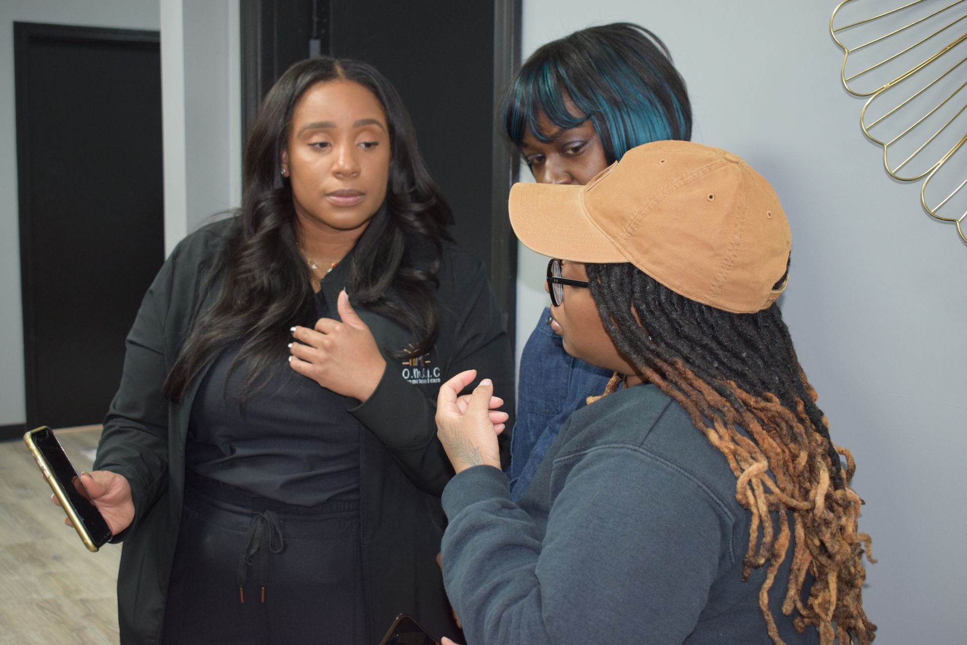 Three people talking indoors, one holding a phone and another wearing a tan cap.