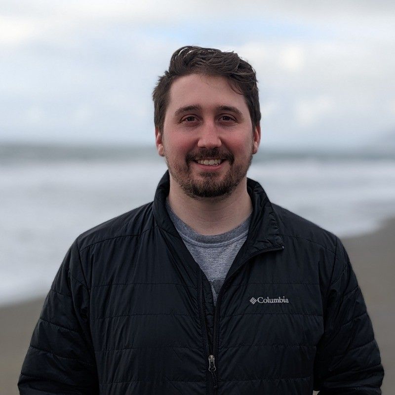 Person standing outdoors in a black jacket, smiling at a cloudy beach