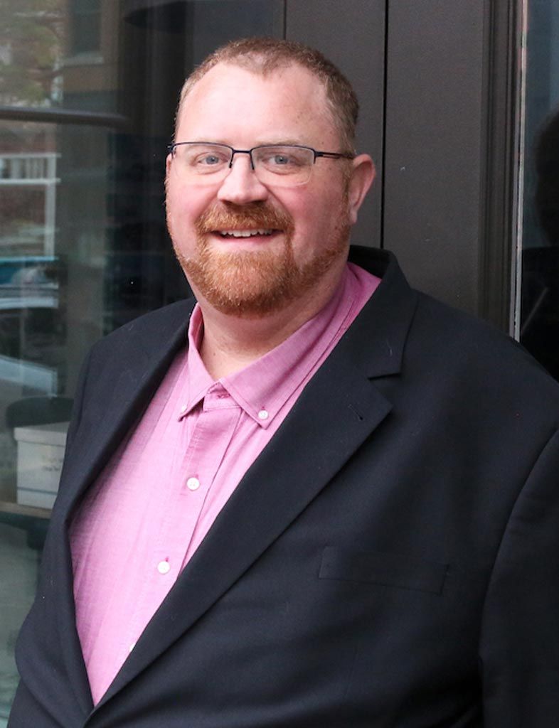 Smiling man in a dark suit jacket and pink shirt standing outdoors by a window