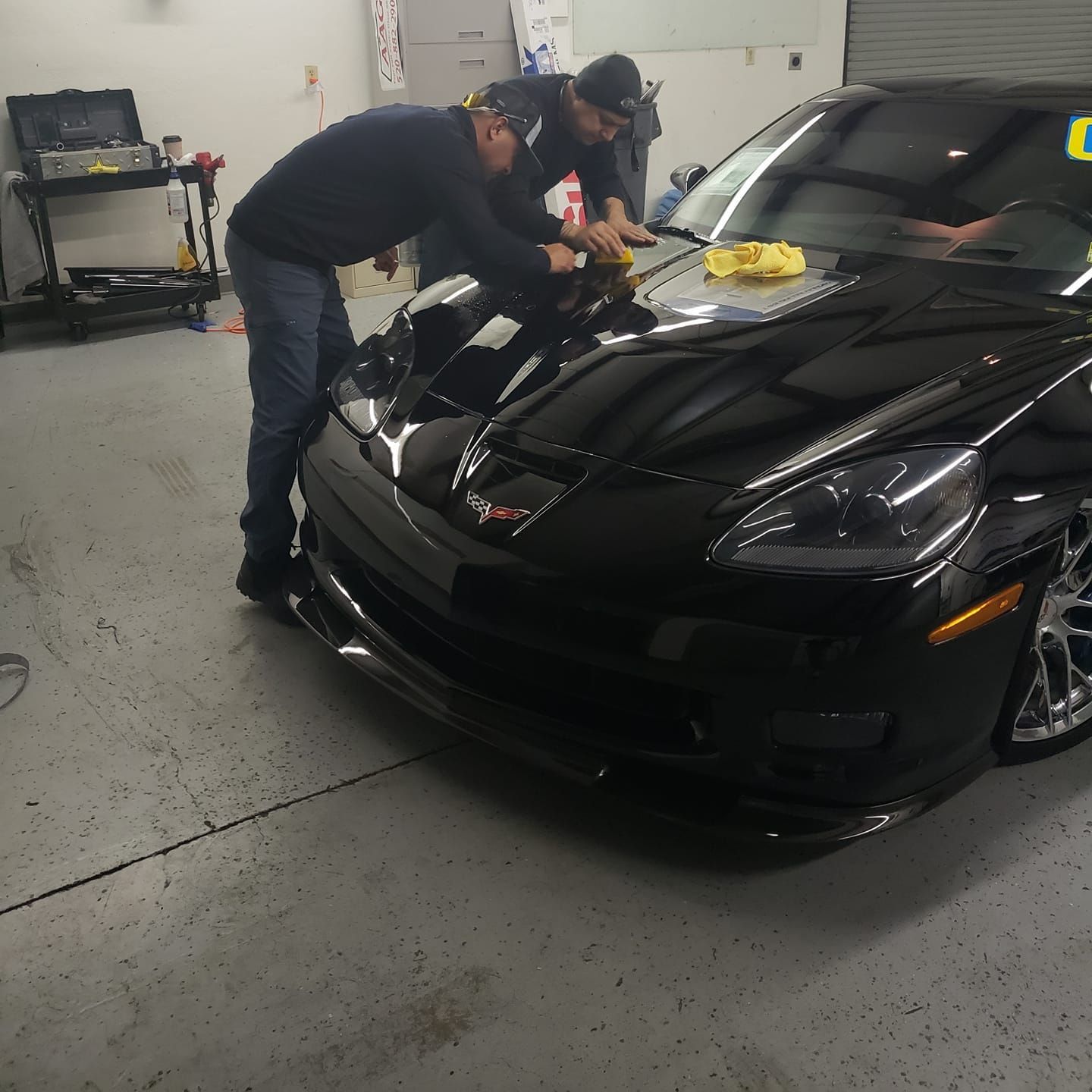 Two men polishing a black Corvette in a garage.