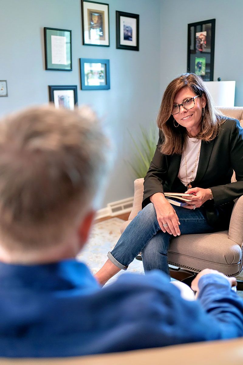Woman in glasses talks with a seated person in a cozy office with framed photos on the wall