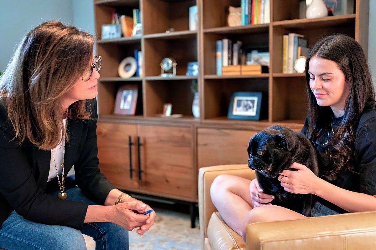 Two women talk in a living room while one holds a small black dog on a couch.