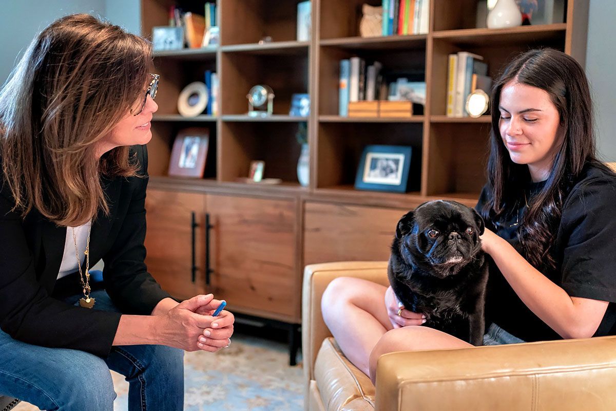 Two women talking in a living room, one holding a small dog on a tan chair.
