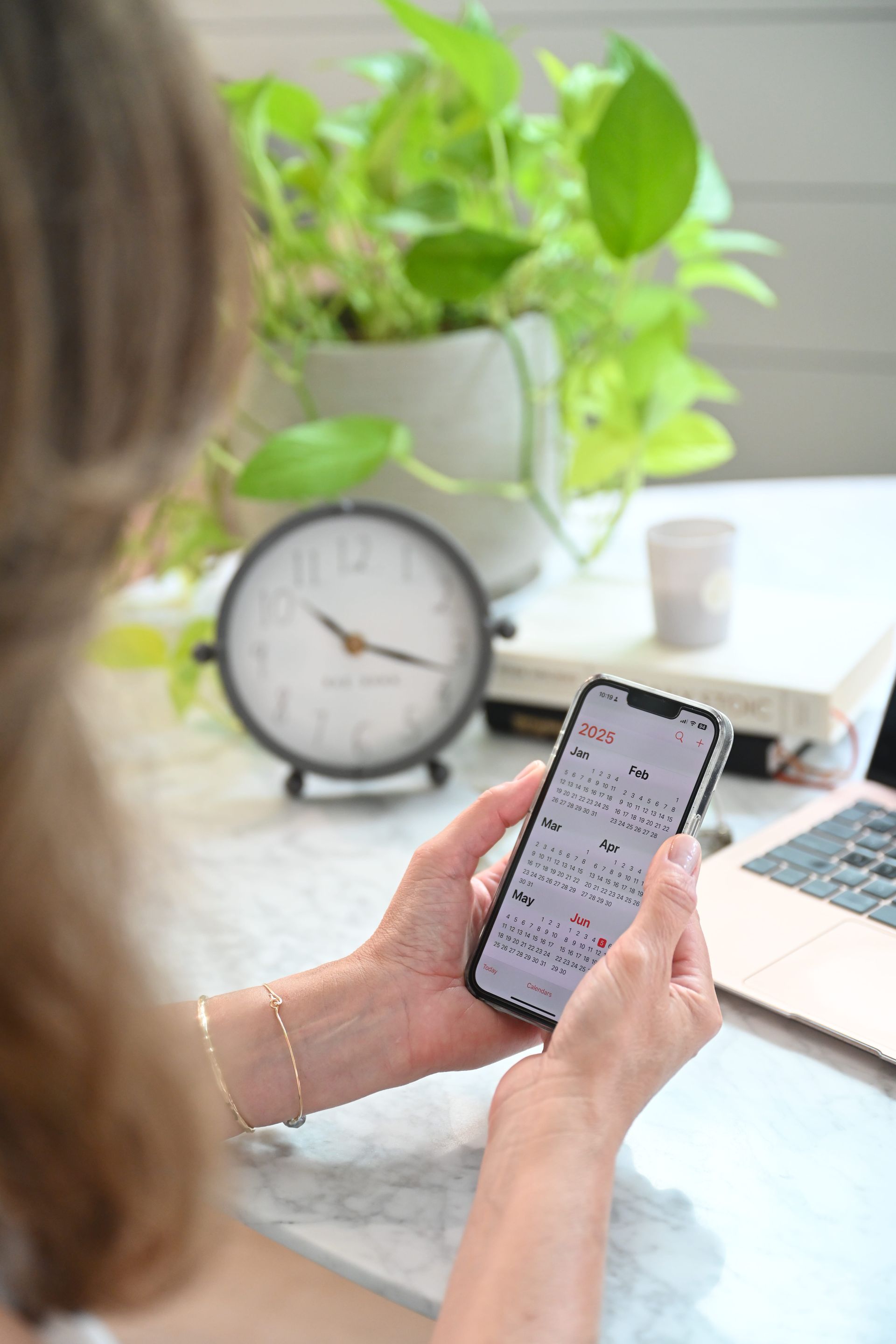 Person holding a smartphone at a desk with a clock, plant, and laptop in the background