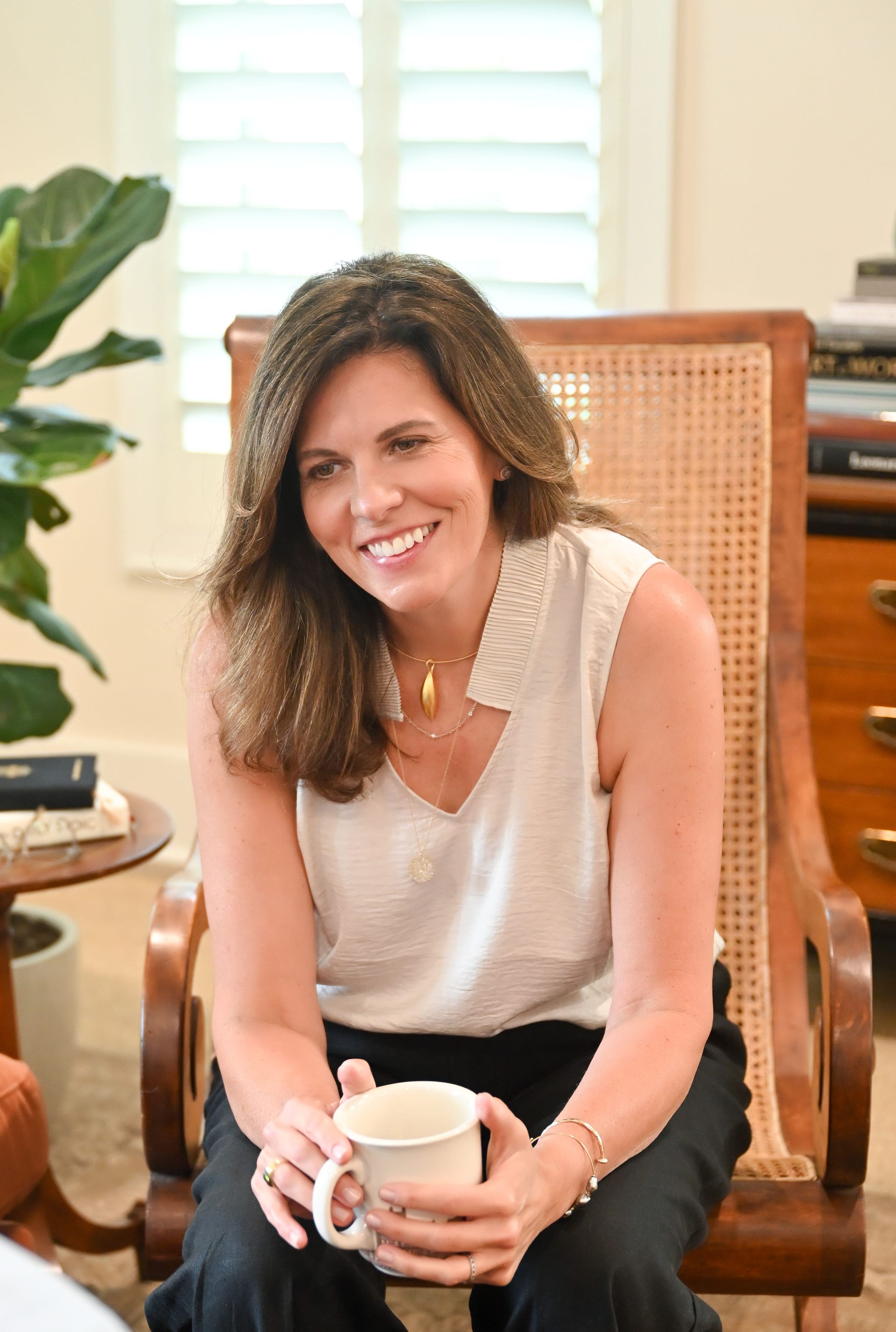 Woman seated in a wicker chair, smiling and holding a mug in a bright room.