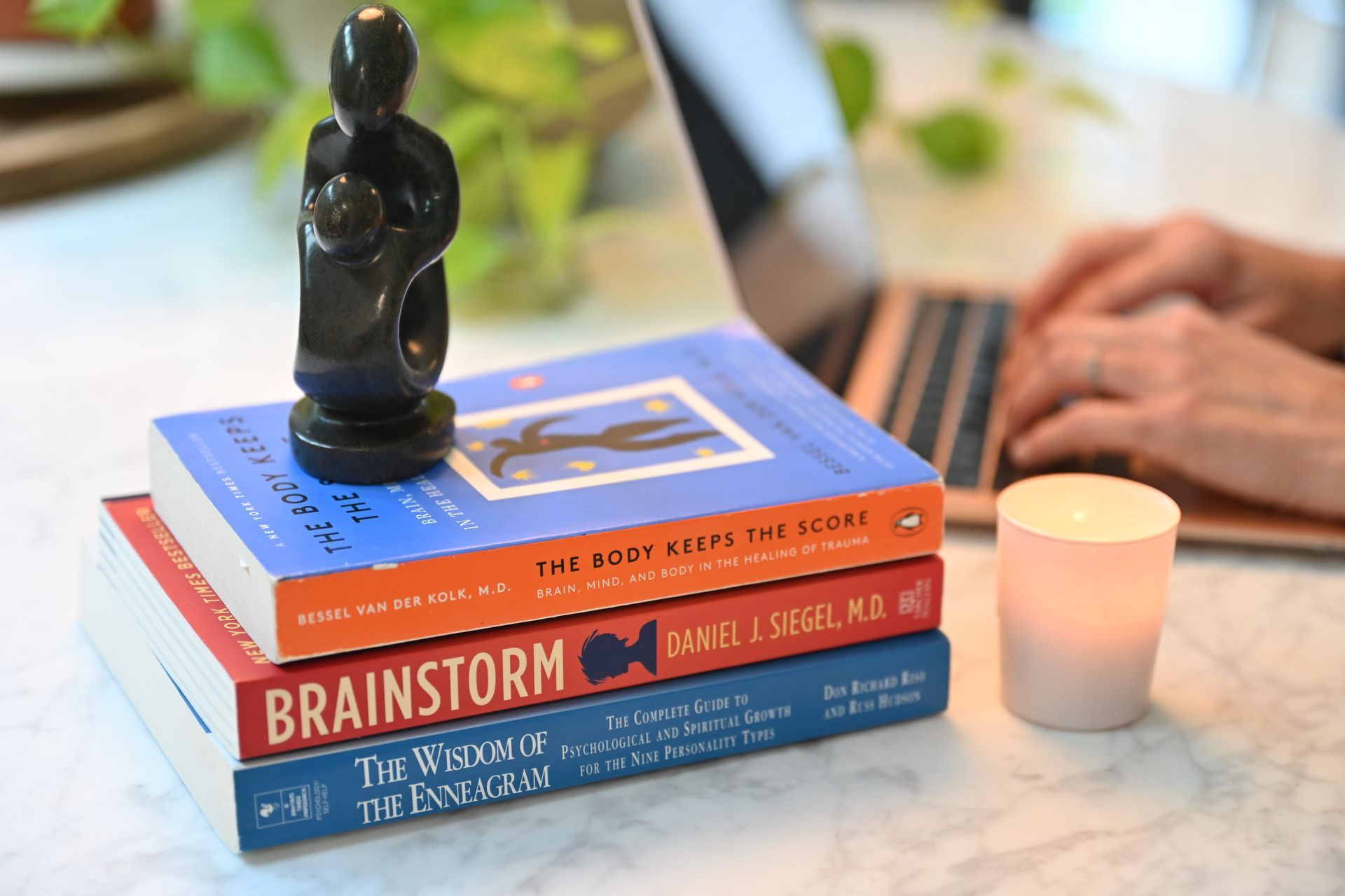 Stack of colorful books with a black figurine on top beside a lit candle and laptop on a table