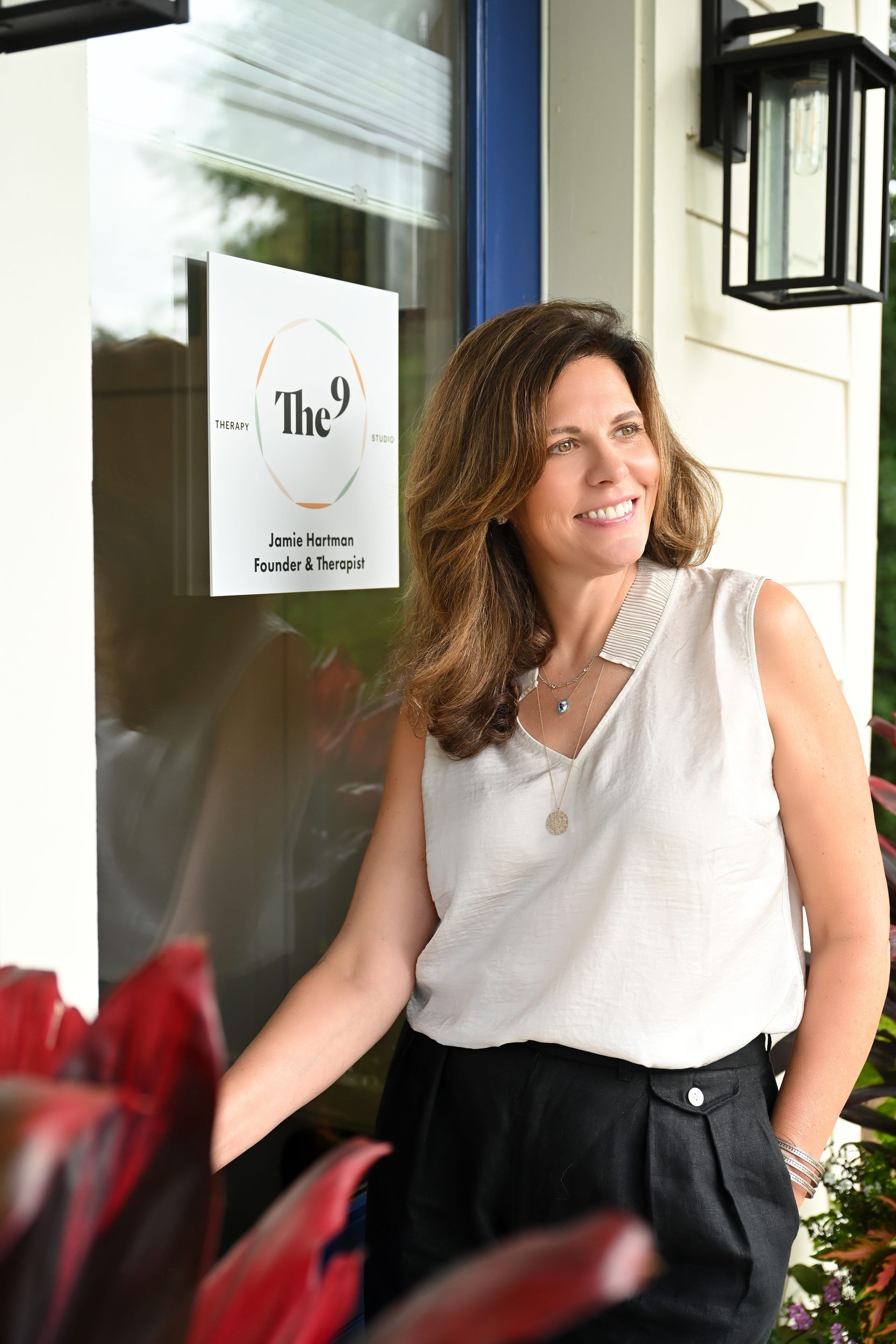 Woman in a sleeveless white blouse stands by a glass door with a sign, smiling outdoors.